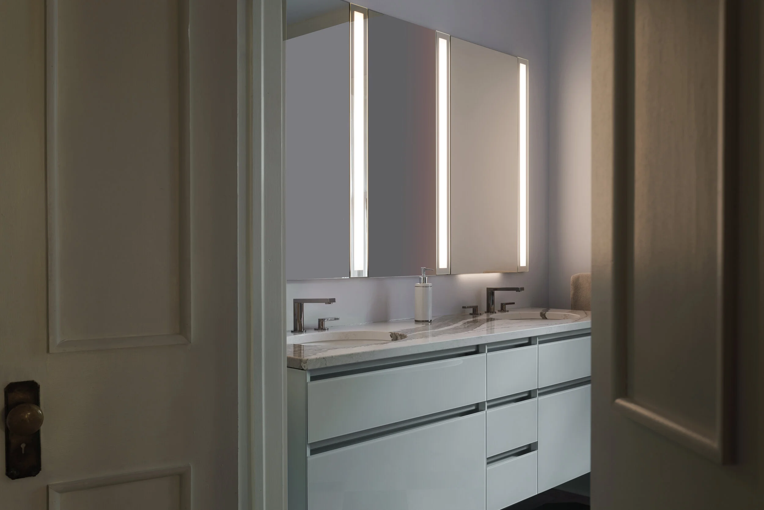 Modern bathroom with double vanity, white cabinets, marble countertop, two sinks, and large illuminated mirror.