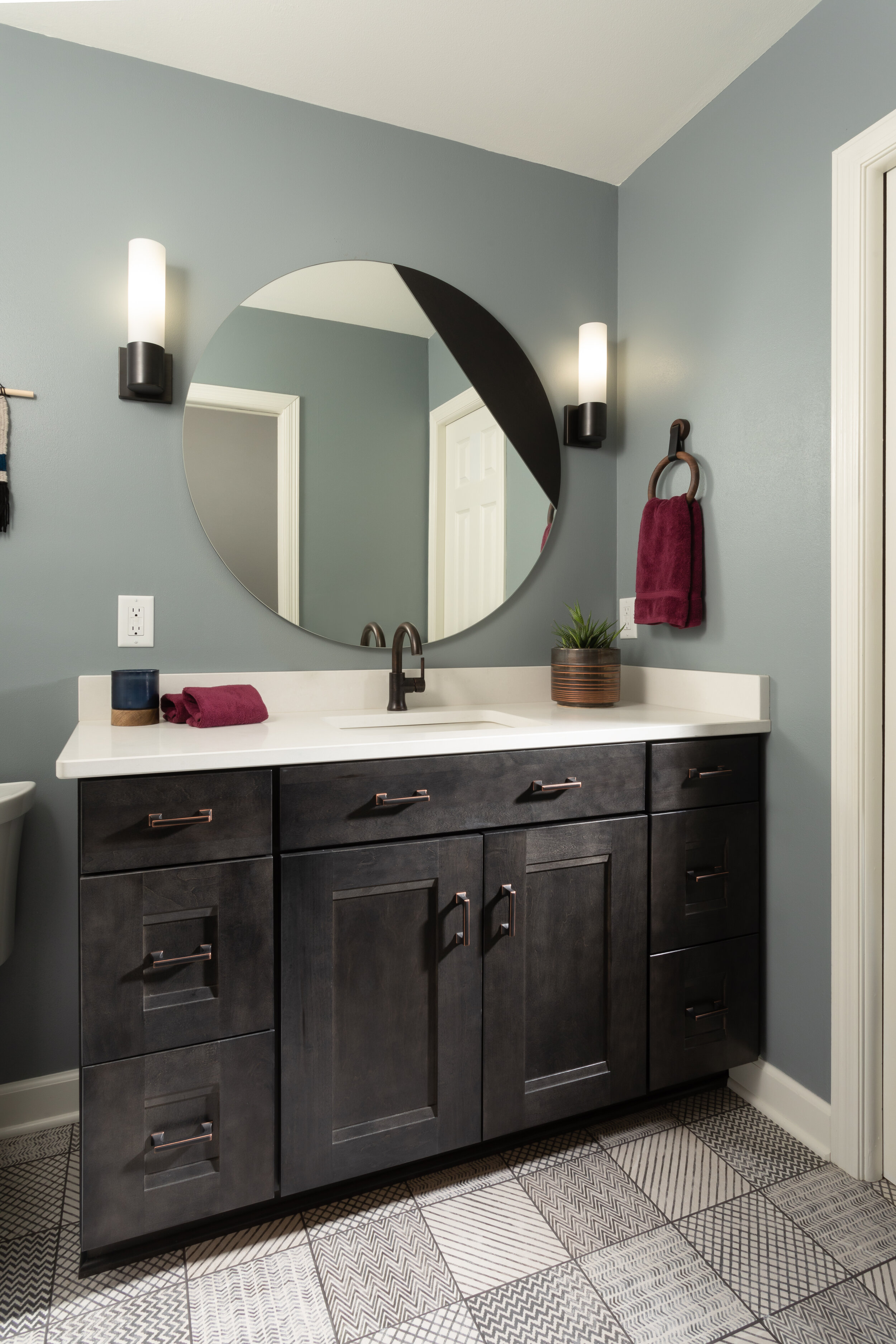 Modern bathroom with a dark gray vanity, white countertop, round mirror, wall-mounted lights, and decorative towels and plant.