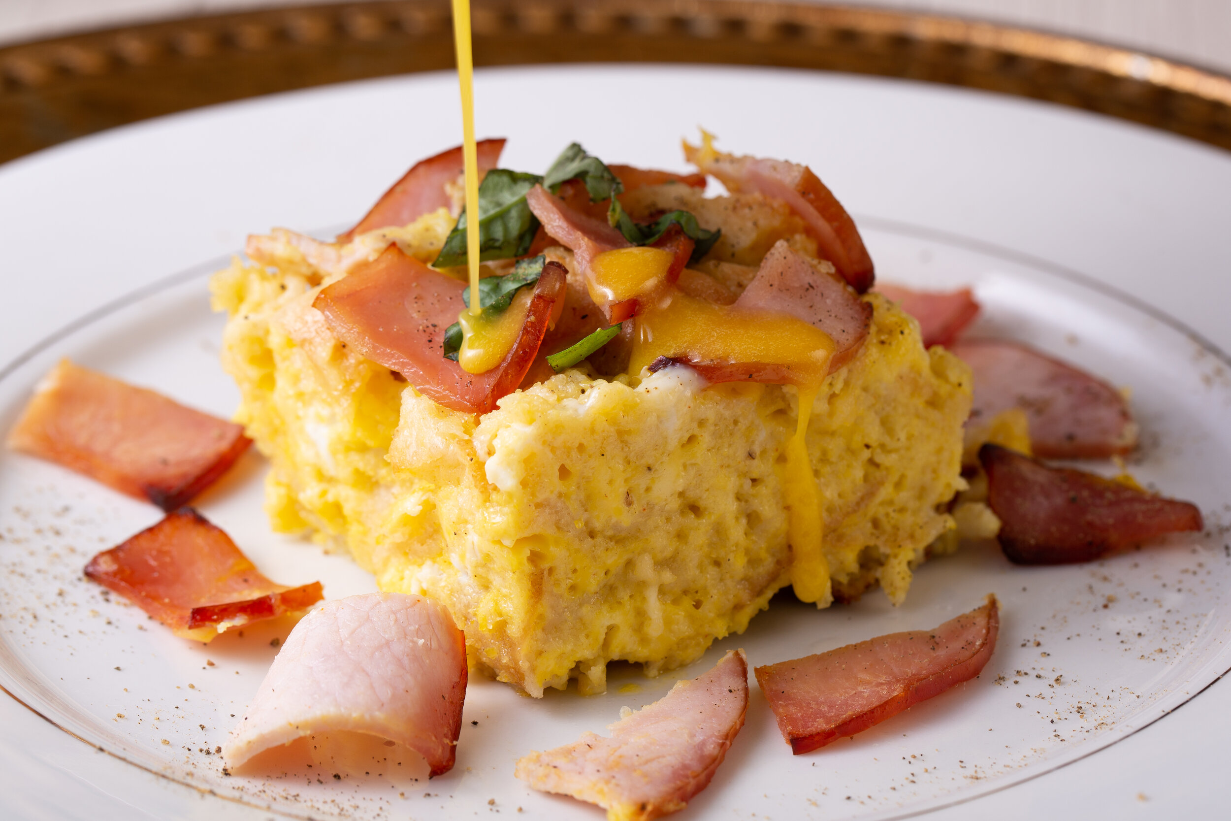A plate of breakfast casserole topped with chopped bacon, cheese, and green herbs, with additional bacon pieces around the casserole on the plate.