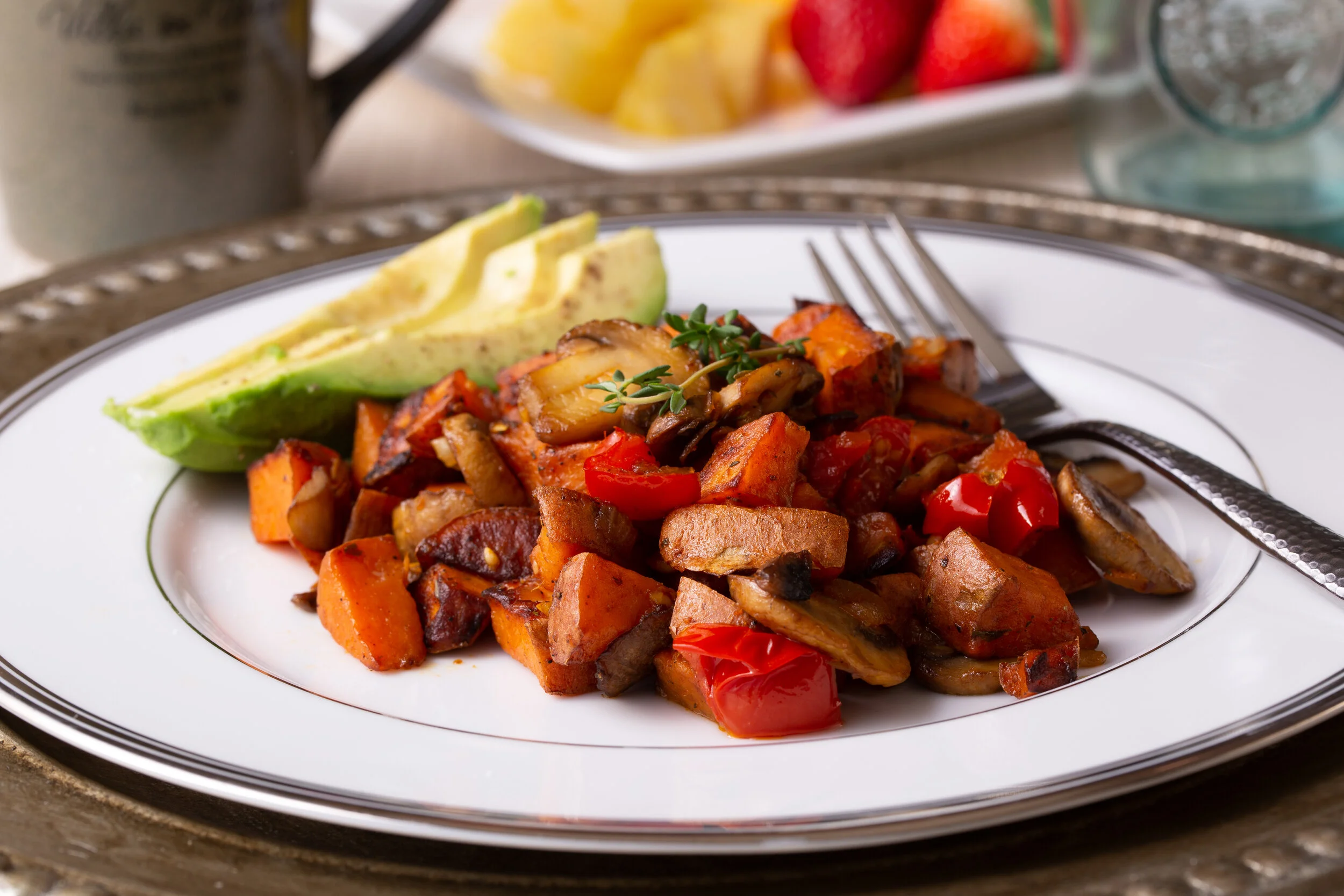 A plate with roasted vegetables, including red bell peppers, mushrooms, carrots, and sweet potatoes, garnished with a sprig of thyme, served with sliced avocado.