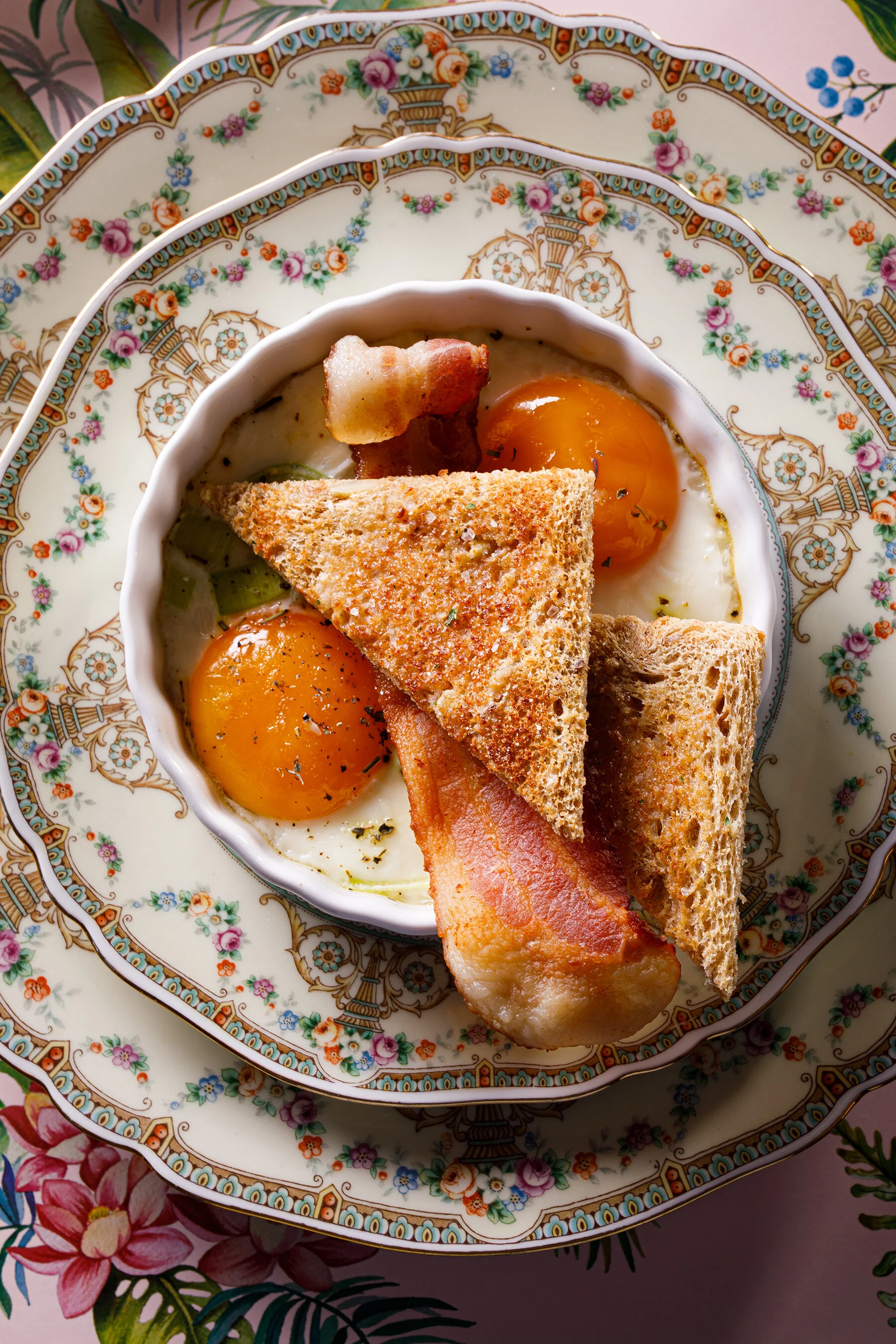 A decorative porcelain plate holding two eggs sunny side up, crispy bacon, toasted bread triangles, and a slice of ham, on a floral-patterned tablecloth.