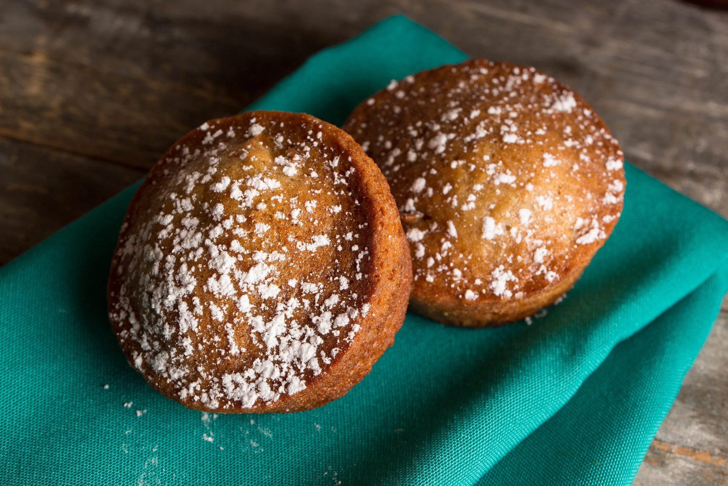 Two muffins with powdered sugar on top, placed on a teal cloth on a wooden surface.