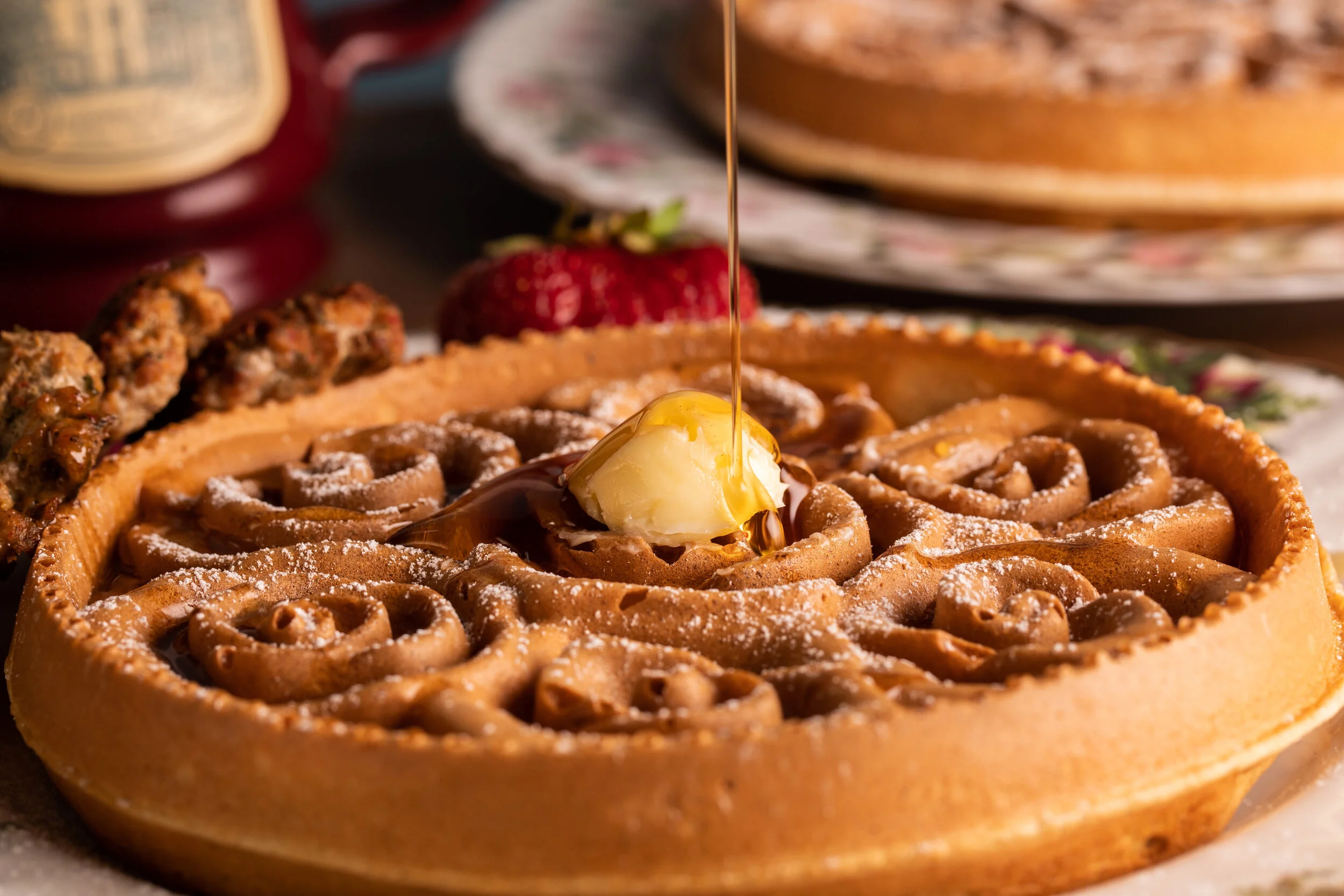 Close-up of a waffle with a pat of melting butter and syrup poured over it, on a decorative plate with strawberries and other breakfast foods in the background.