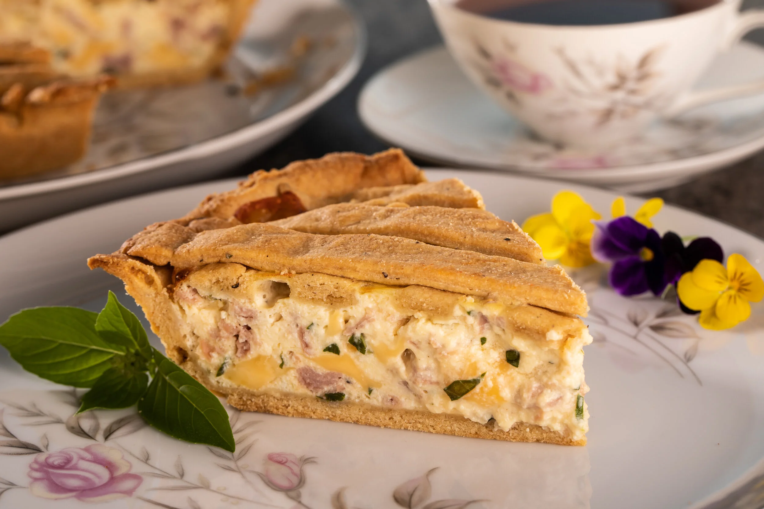 Slice of homemade savory pie with a flaky crust, filled with eggs, ham, cheese, and green herbs, garnished with fresh basil and edible flowers on a floral china plate.