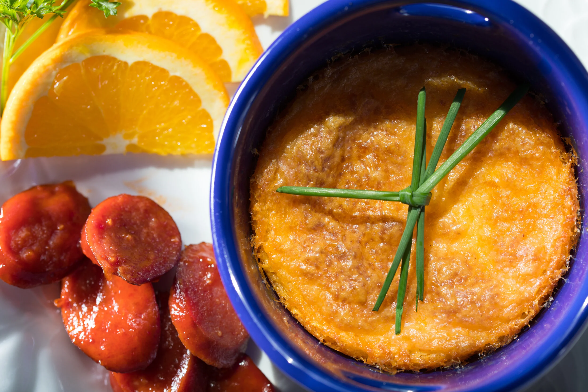 Baked macaroni and cheese in a blue bowl garnished with chives, served with orange slices and smoked sausage on a white plate.