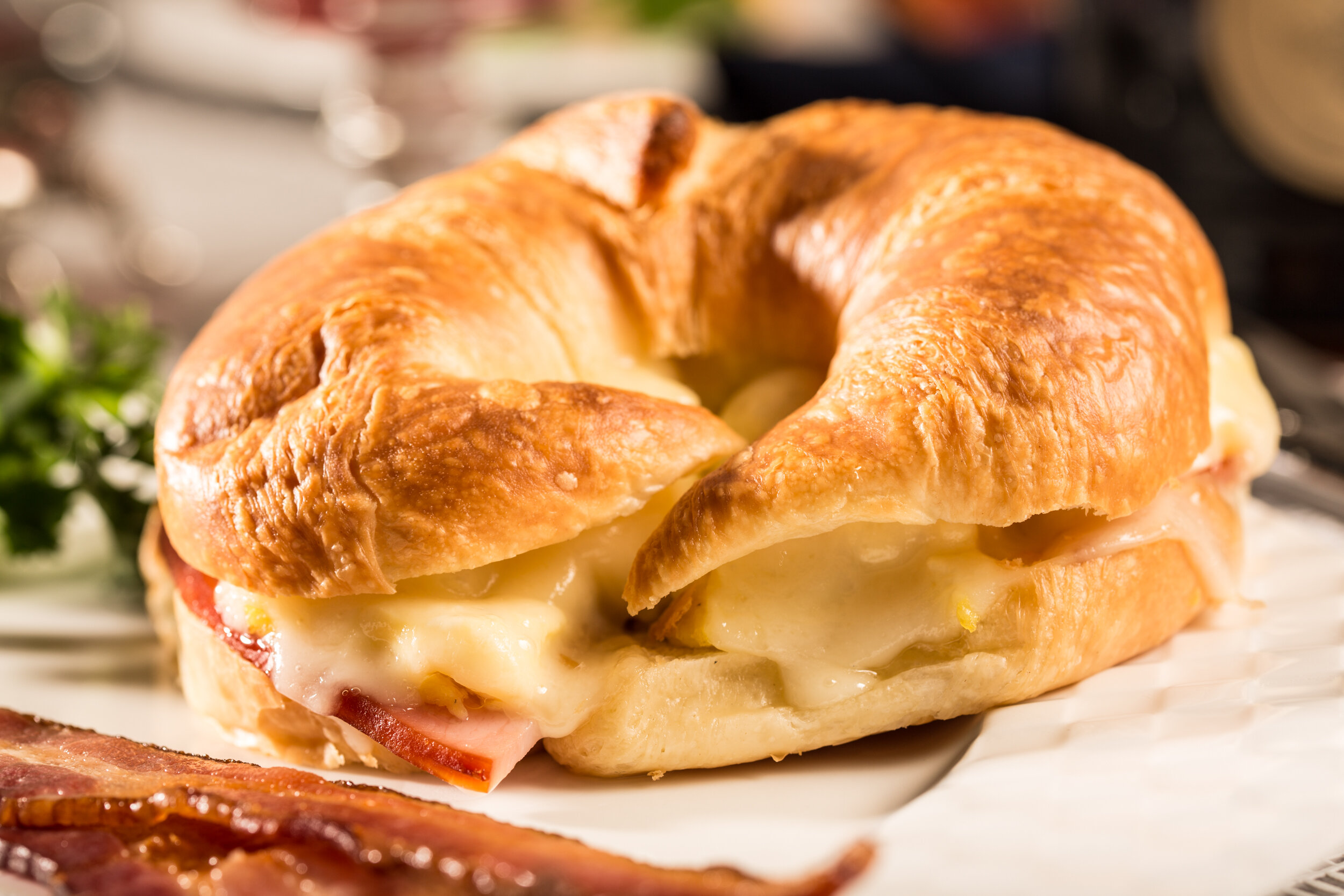 Close-up of a croissant sandwich filled with melted cheese, ham, and possibly eggs, on a white plate.