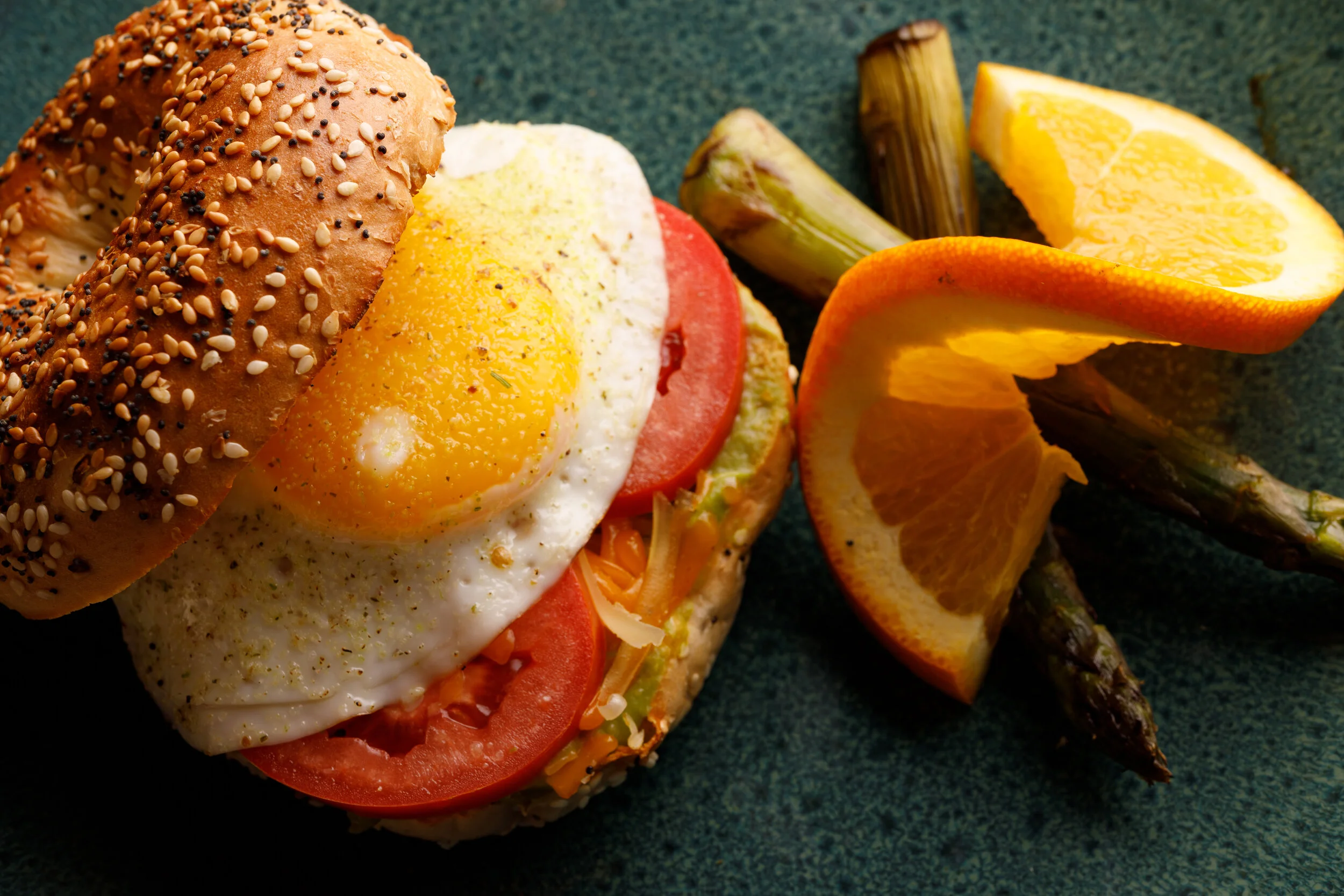 Breakfast sandwich with a fried egg, tomato slices, cheese, and a multigrain bagel on a dark green plate, with orange slices, asparagus, and lemon wedges beside it.