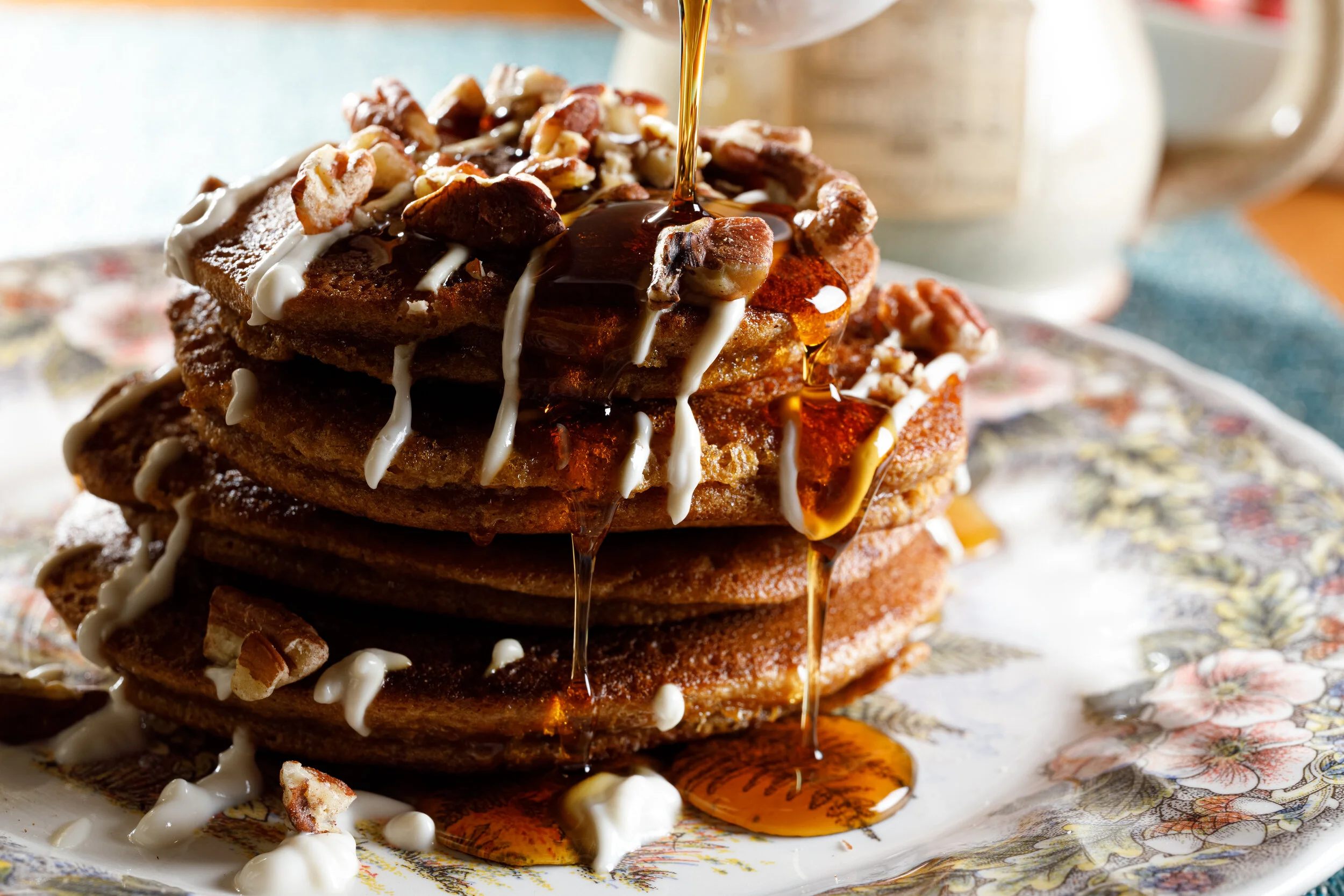 Stack of four thick, fluffy pancakes topped with chopped nuts, drizzled with white icing, and syrup pouring over the top, on a decorative plate with a teapot and cup in the background.