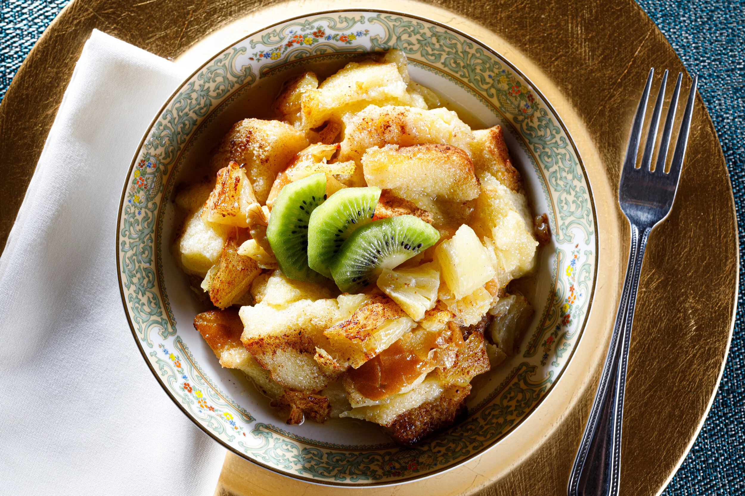 A bowl of bread pudding garnished with sliced kiwi fruit, placed on a gold-rimmed plate with a fork and a white napkin nearby.