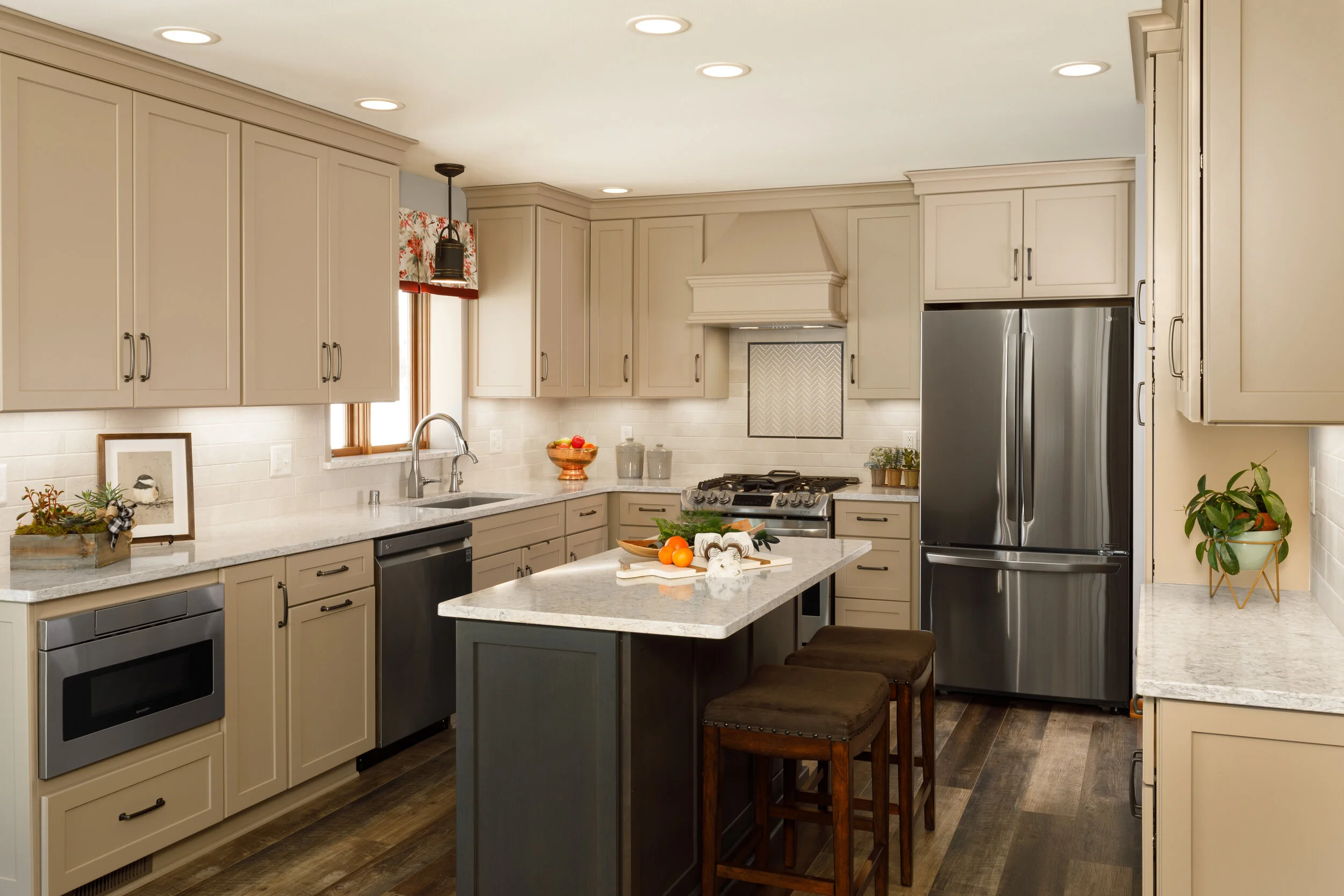 Modern kitchen with beige cabinets, stainless steel refrigerator, microwave, dishwasher, and oven. Kitchen island with stools, white countertops, and decorative plants and framed picture.