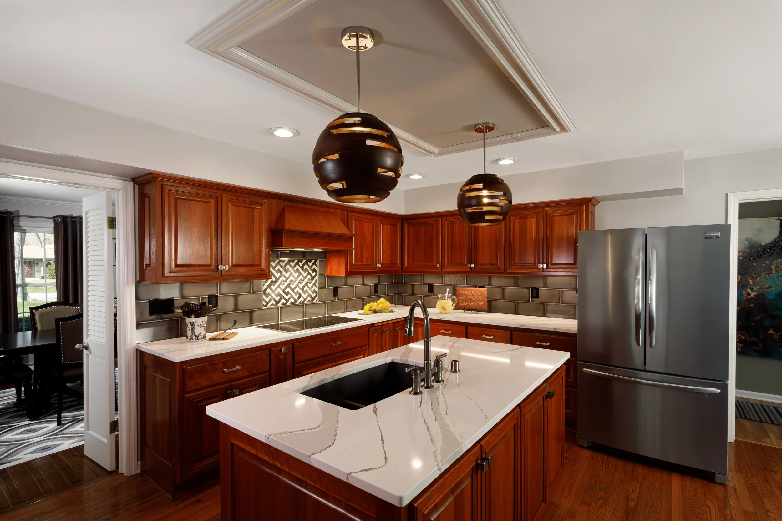 Modern kitchen with wooden cabinets, white marble countertops, stainless steel refrigerator, and black pendant lights, view into adjacent dining area.