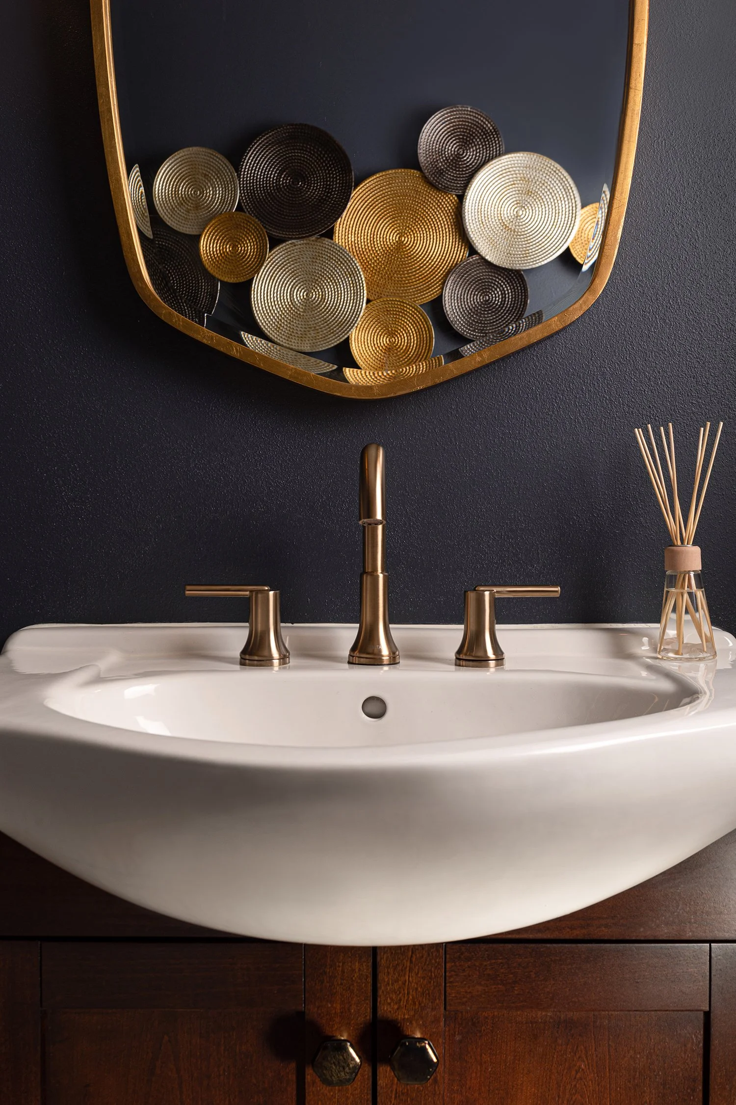 A bathroom sink with two handles, a central faucet, mounted on a wooden vanity against a dark wall. Above the sink, a mirror with a gold frame reflects decorative circular gold, black, and silver objects.