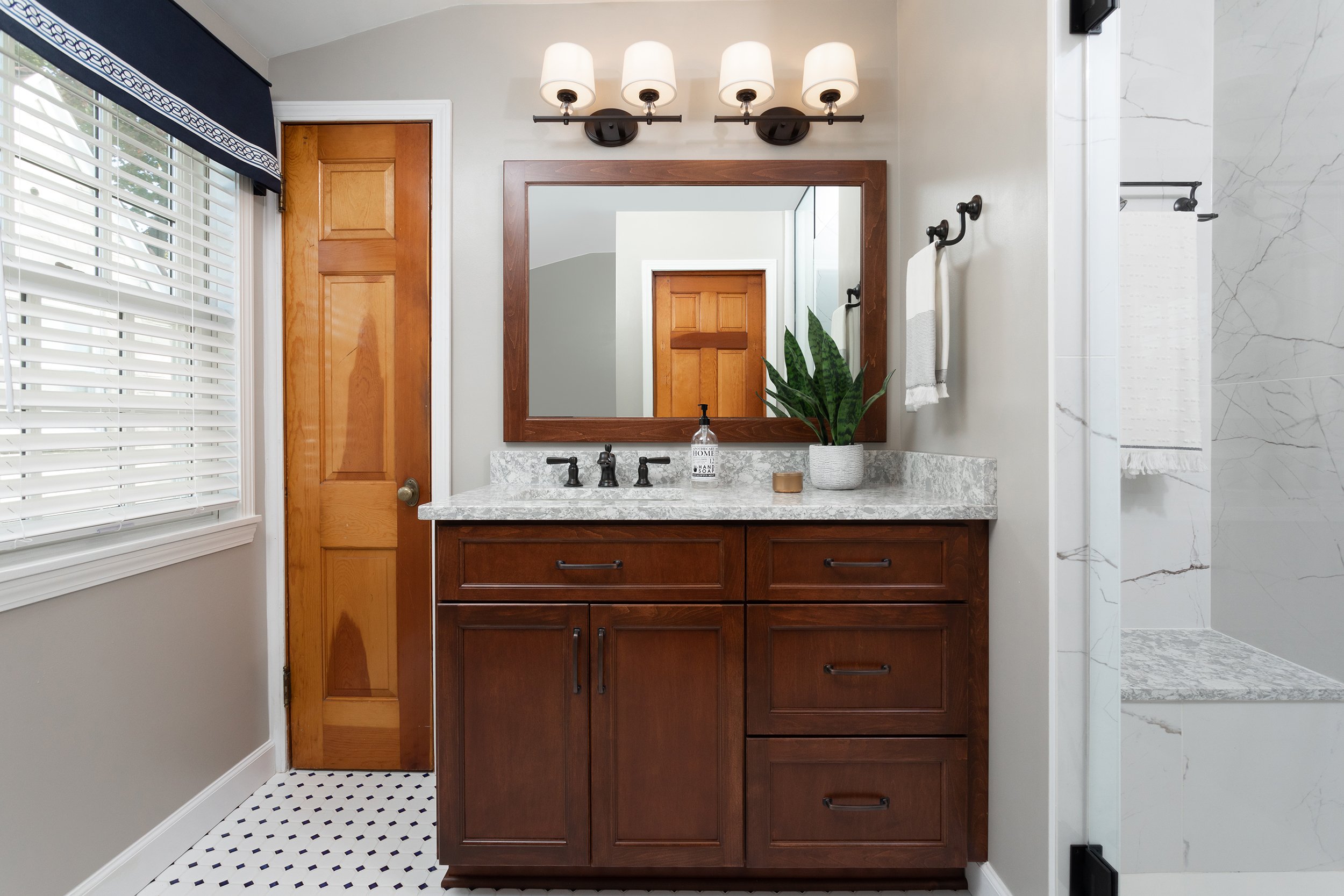 Bathroom vanity with a large mirror, wooden cabinet, marble countertop, potted plant, soap dispenser, and towels. There is a window with blinds and a wooden door. Part of a marble shower is visible on the right side.