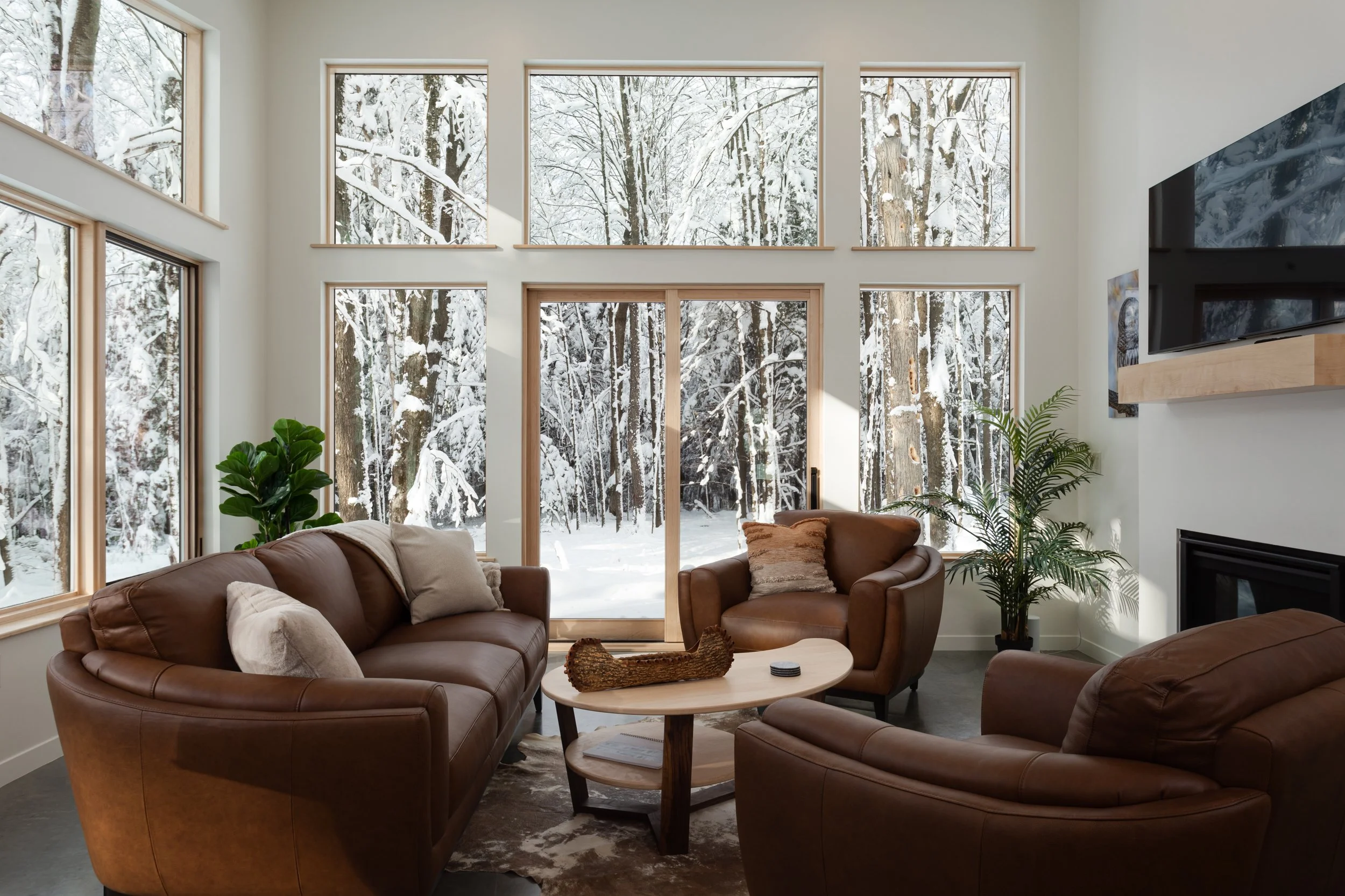 Living room with large windows showing snowy forest outside, brown leather sofas, a beige armchair, a wooden coffee table, indoor plants, and a fireplace.