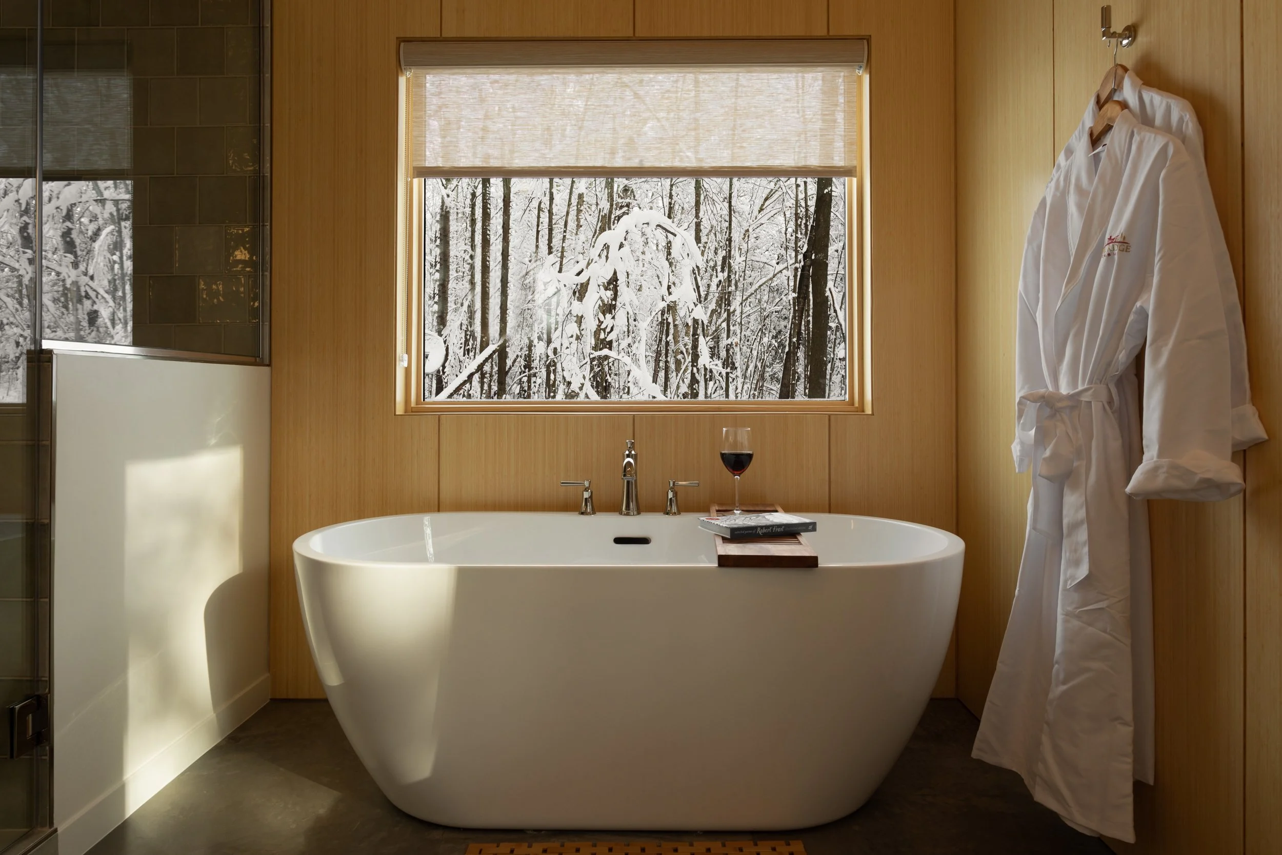 A cozy bathroom featuring a free-standing bathtub beneath a window with a snowy winter landscape outside, with a robe hanging on the wall and a glass of red wine on a tray beside the tub.
