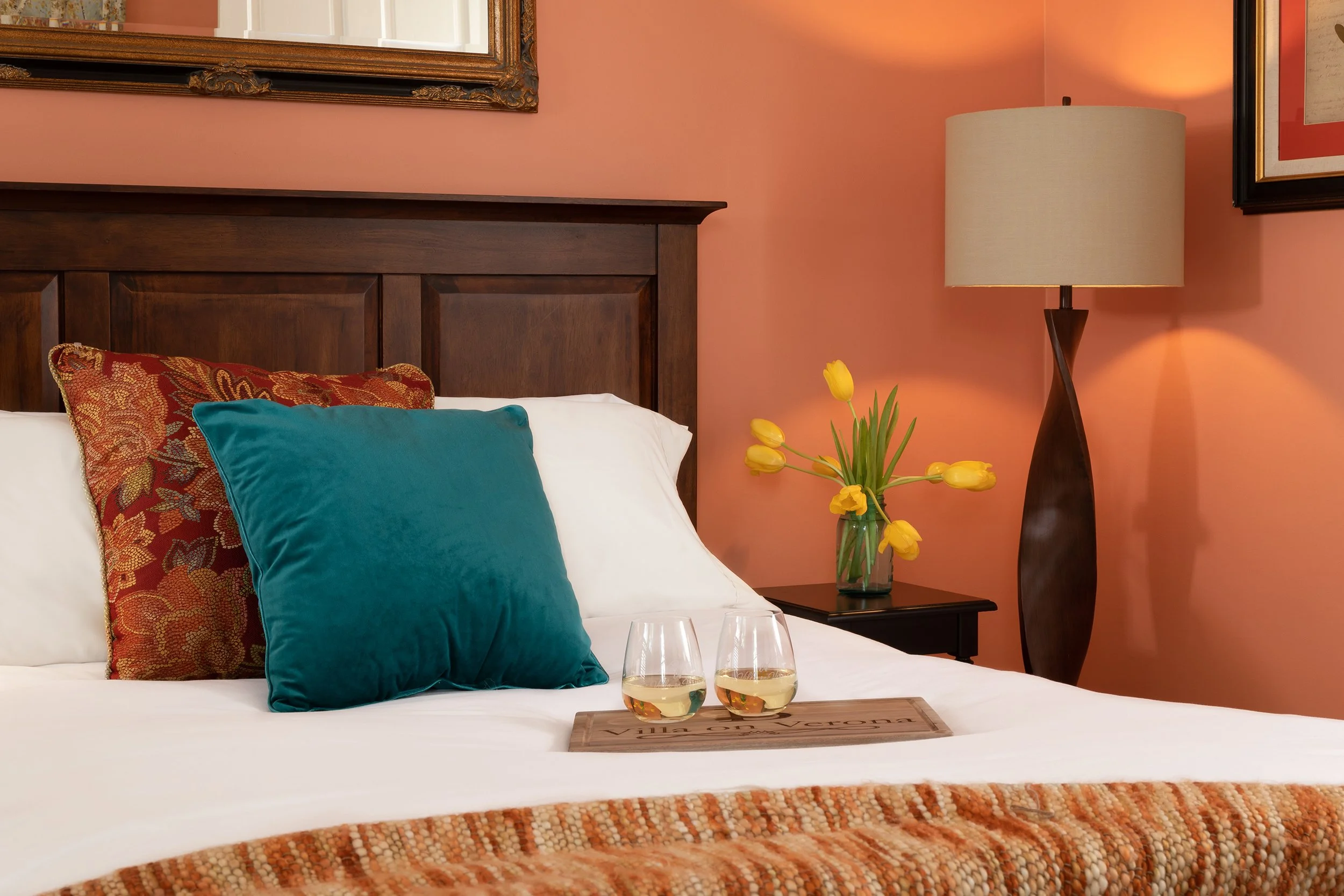 A neatly made bed with colorful pillows, a tray with two glasses of white wine, a side table with a vase of yellow tulips, and a tall table lamp against a peach-colored wall in a cozy bedroom.