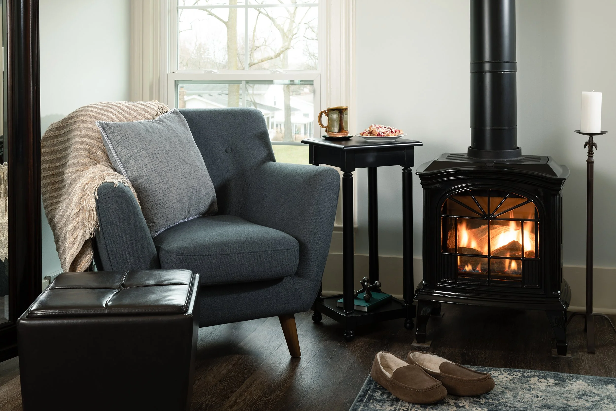 Living room corner with a blue armchair, a black side table with a mug and snacks, a black fireplace with a fire, a white candle on a brass stand, a pair of slippers on a rug, and a window showing a tree outside.
