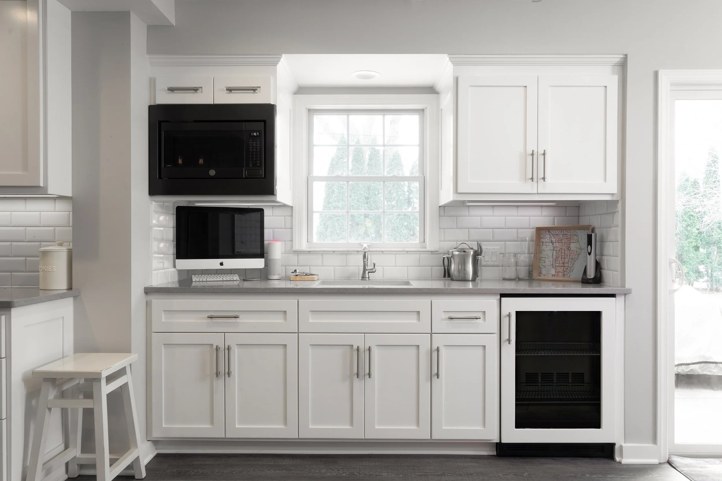 Kitchen with white cabinets, gray countertop, window above sink, black microwave, iMac computer, wine cooler, and small barstool on left.