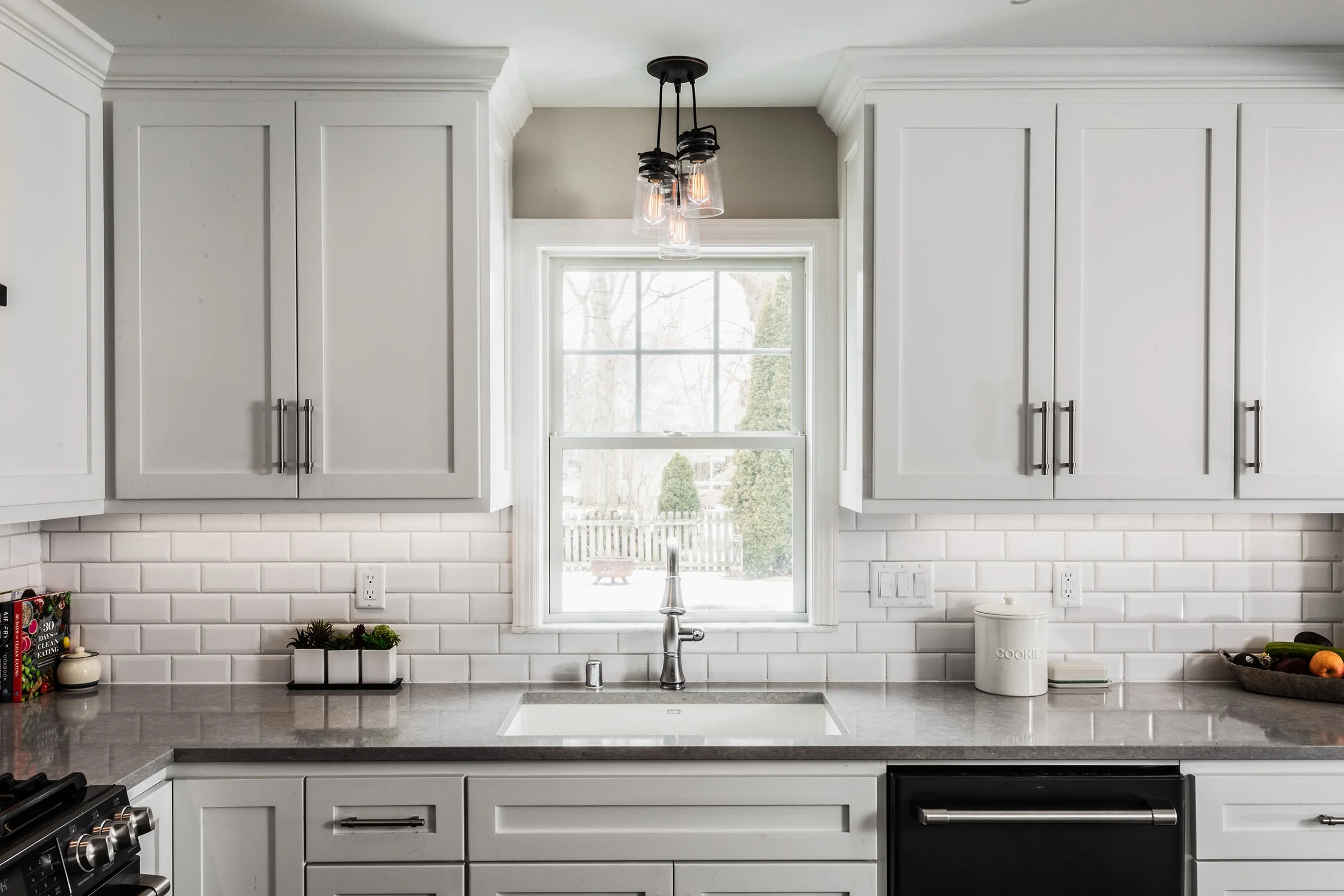 Kitchen with white cabinets, gray countertop, and subway tile backsplash, featuring a window above the sink, a black dishwasher, and a modern hanging light fixture.