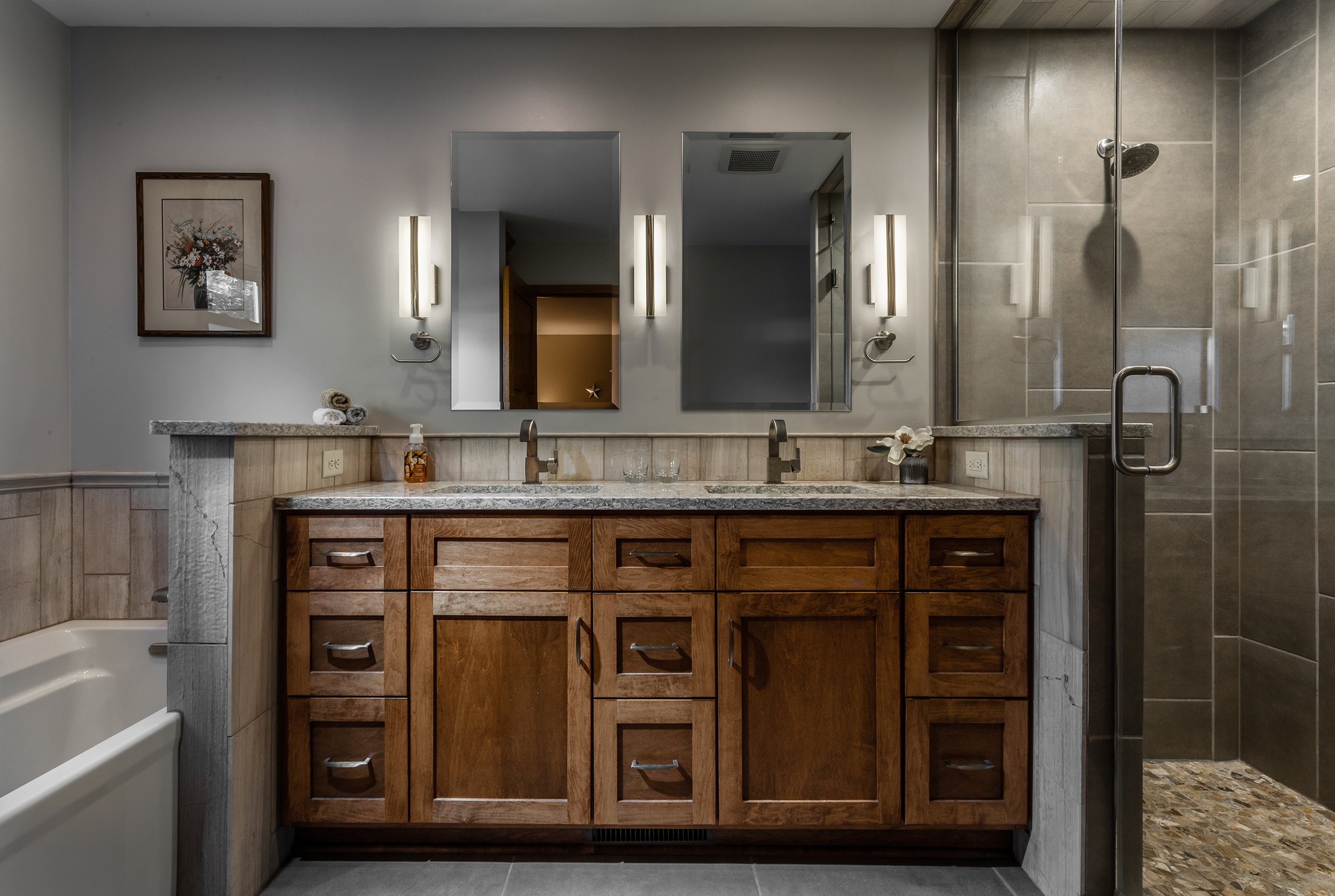 Bathroom with double vanity, wooden cabinets, granite countertop, two mirrors, wall-mounted lights, and a glass-enclosed shower.