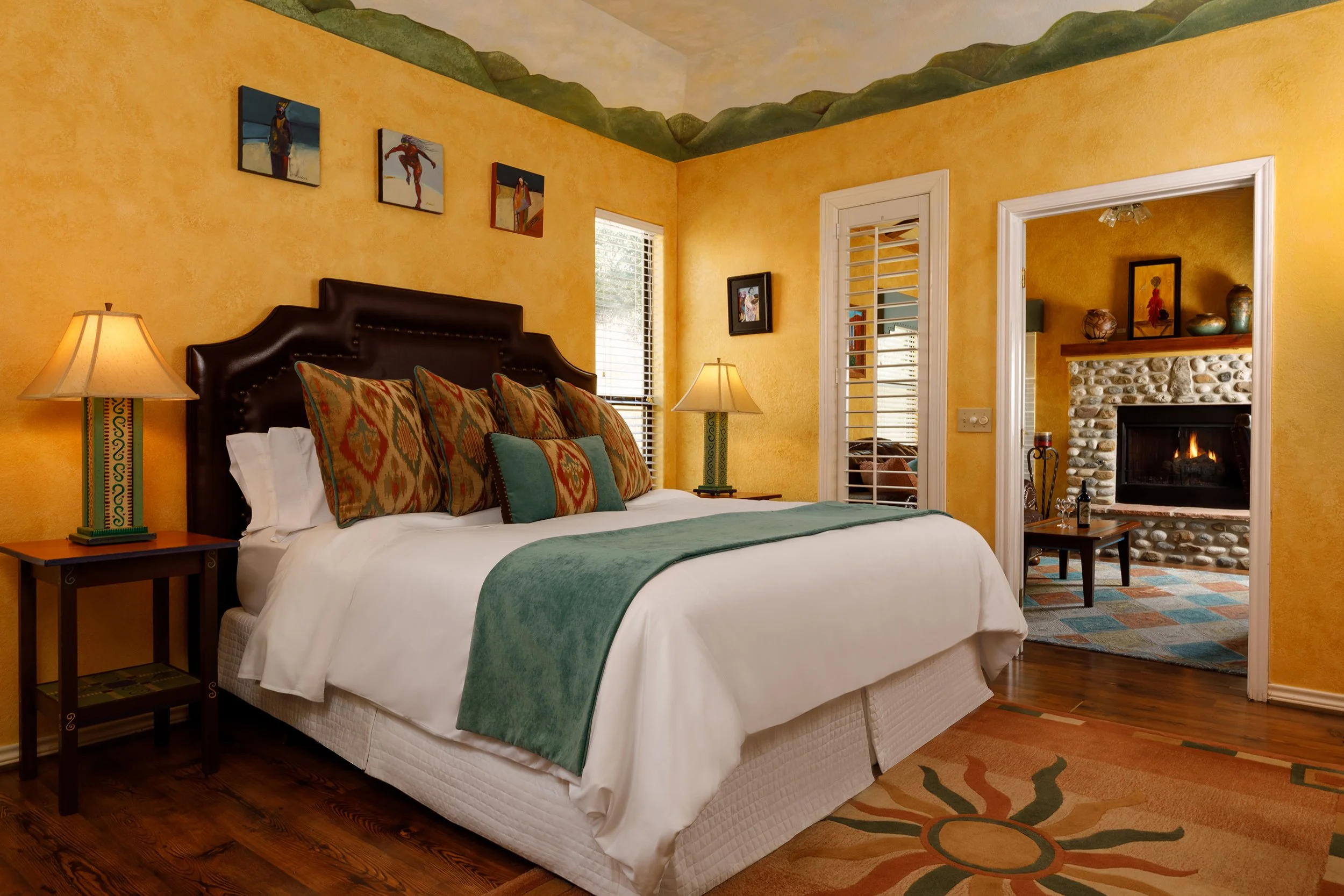 A cozy bedroom with yellow textured walls, dark wooden flooring, a large bed with white bedding, colorful patterned pillows, a green throw blanket, and a dark headboard. There are two bedside tables with lamps. A window with blinds and a doorway lead