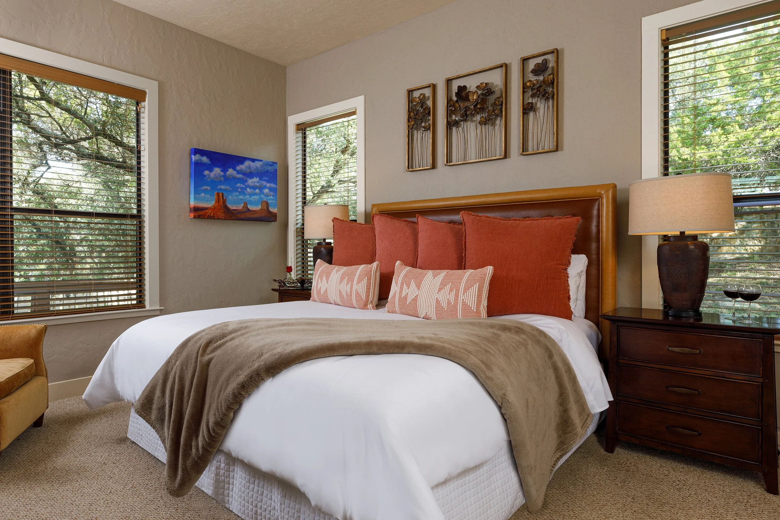 Cozy bedroom with a large bed, multiple orange and patterned pillows, beige blanket, wooden nightstand with a table lamp, wall art, and windows with green trees outside.