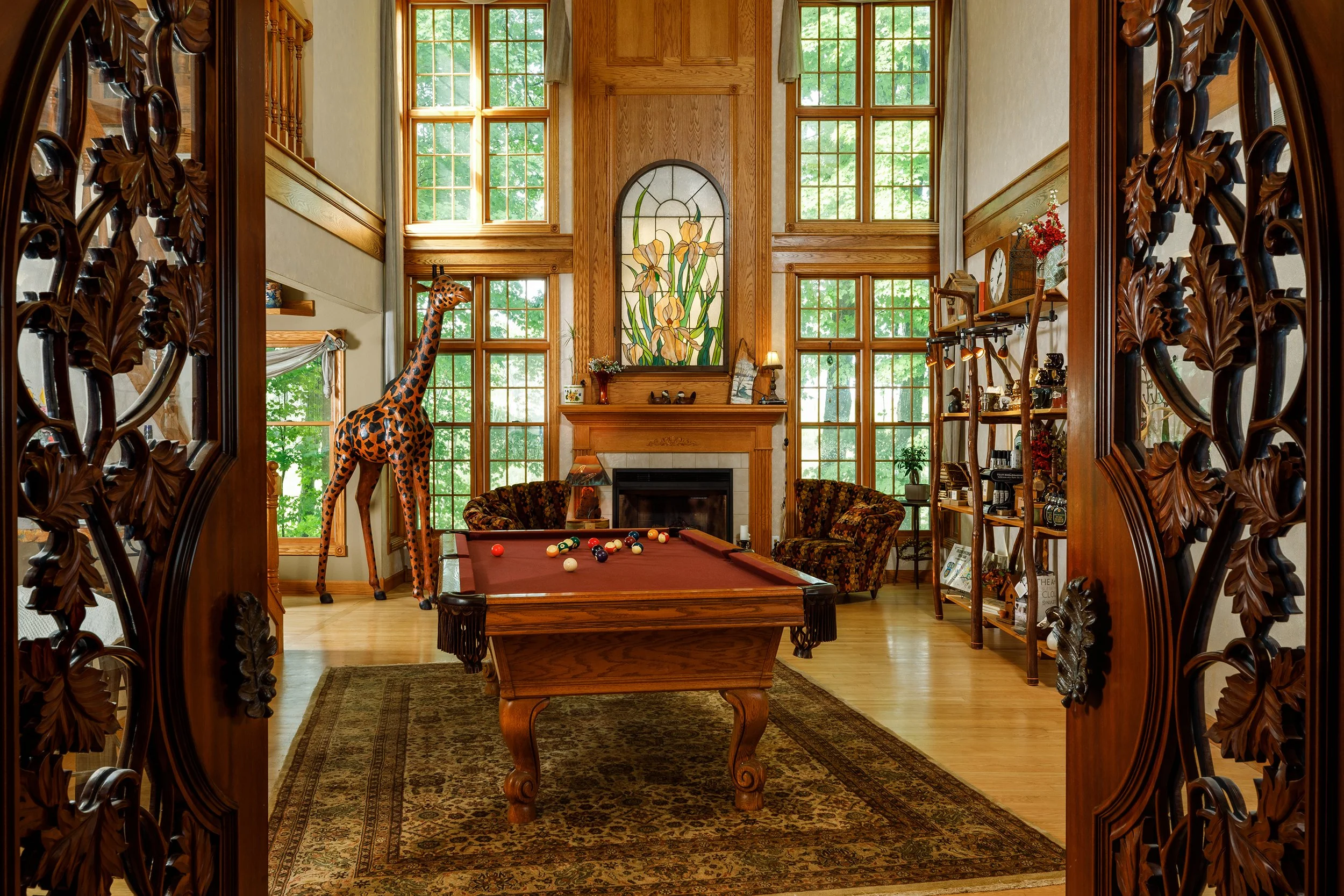Interior of a living room with tall wooden windows, a fireplace, a stained glass art piece above the fireplace, a pool table with balls, a giraffe statue near the windows, and shelves with decorative items and books.