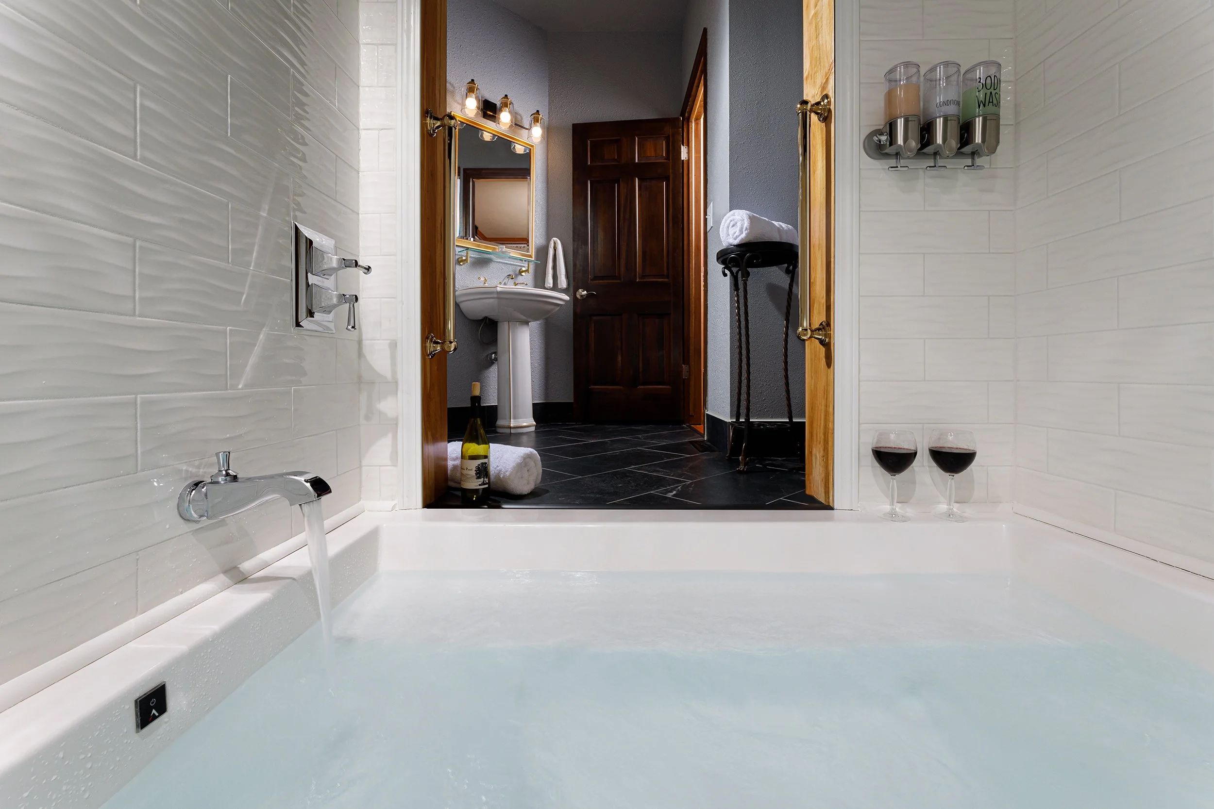 Private bathroom with bathtub in foreground, white tiled walls, and a view into a separate room with dark flooring, a mirror, sink, towels, wine, and glasses of red wine.