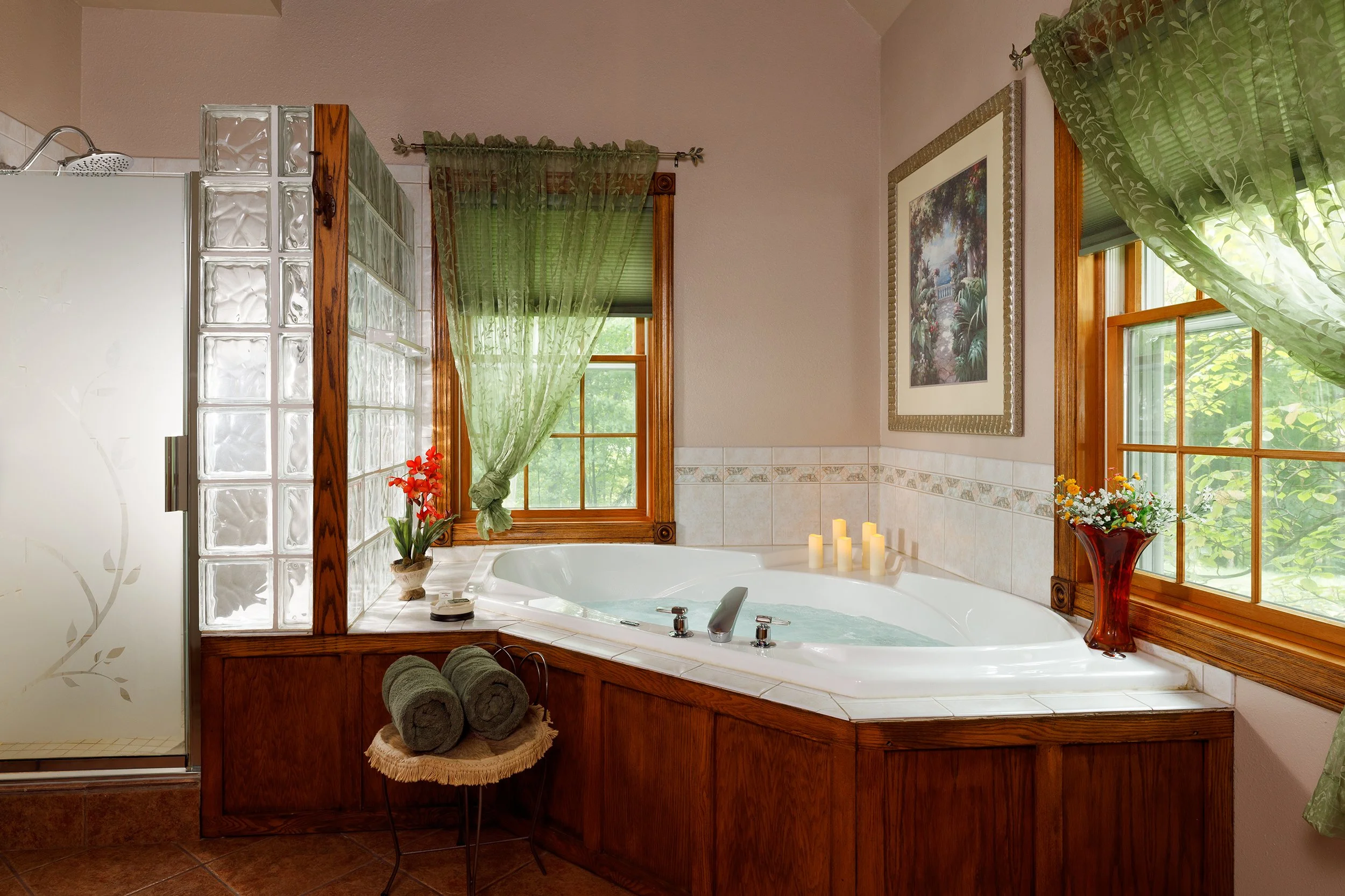 Bathroom with a corner white bathtub, surrounded by wooden paneling, green curtains on windows, candles on the ledge, and decorative flowers.