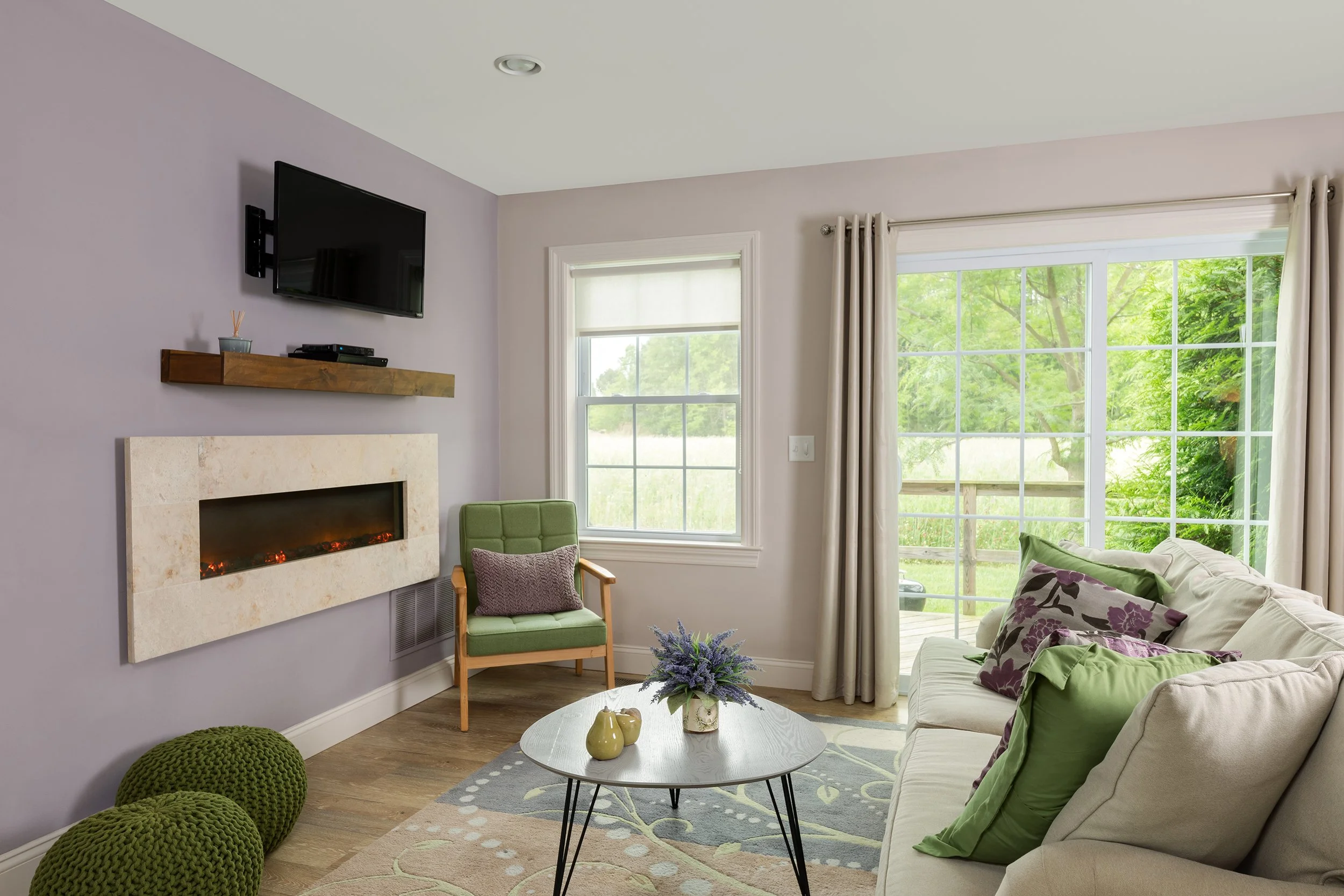 Living room with purple wall, electric fireplace, green armchair, cream sofa with purple and green pillows, wooden coffee table with flowers, window and sliding door with cream curtains, outside greenery visible.