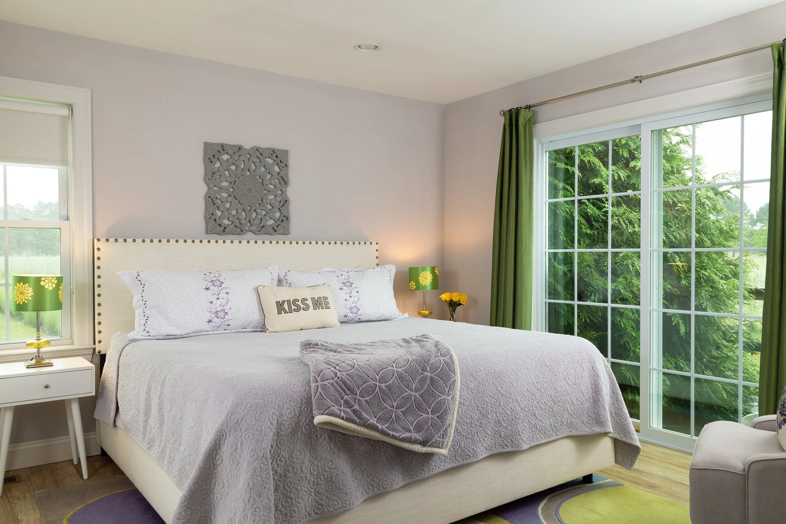 A neatly made bedroom with a large bed, purple and white bedding, and a pillow with 'KISS ME' text. There are nightstands with green lamps, a window with green curtains, and a sliding glass door showing green trees outside.