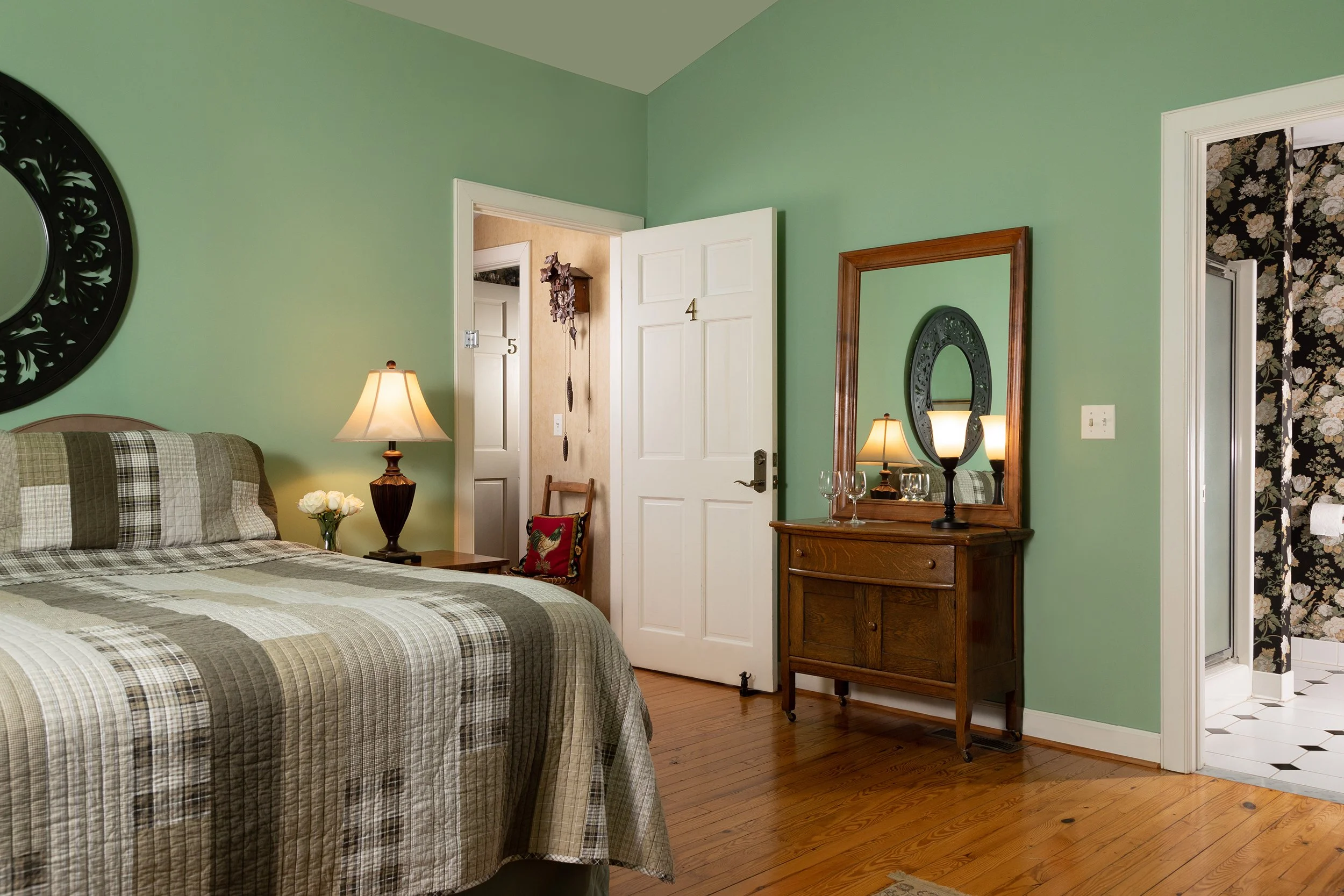 A bedroom with green walls, a bed with a plaid bedspread, a nightstand with a lamp and flowers, a wooden dresser with a mirror and lamps, and an open doorway leading to a bathroom with floral wallpaper.