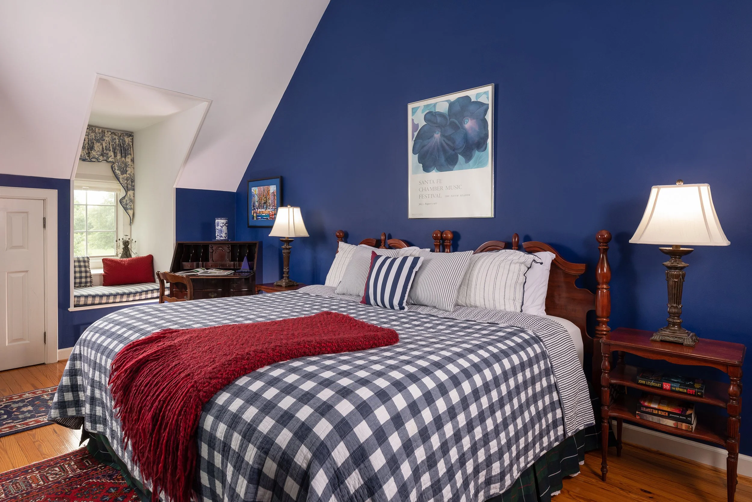 A cozy bedroom with a blue accent wall, wooden bed with a checkered black and white bedspread, decorative pillows, and a red throw blanket. There are two matching bedside lamps, and art posters on the walls. A small nook with a window seat and patter