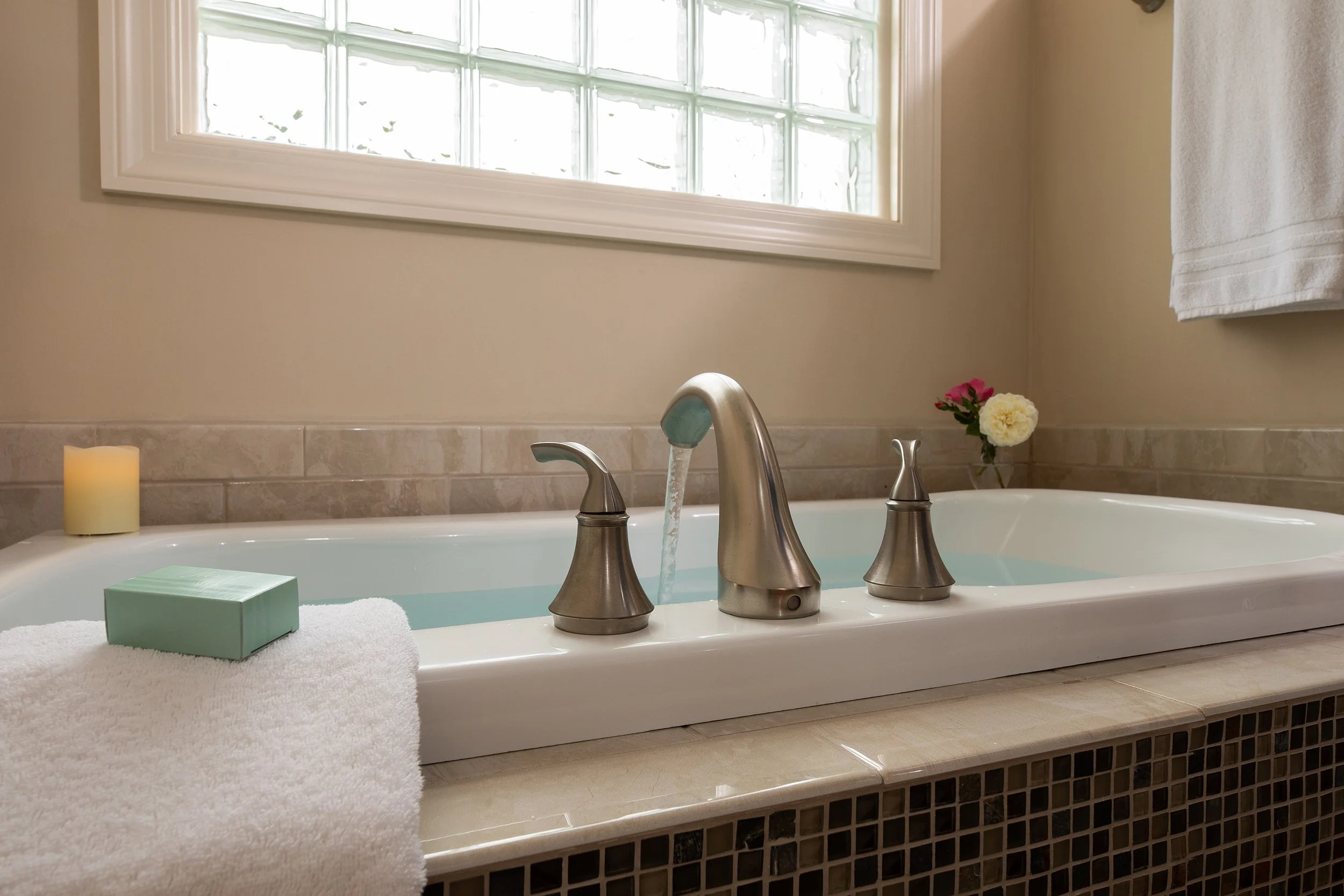 Bathroom bathtub with silver faucet running water, a white towel, a bar of soap, a candle, a flower in a small vase, and a window with glass blocks.