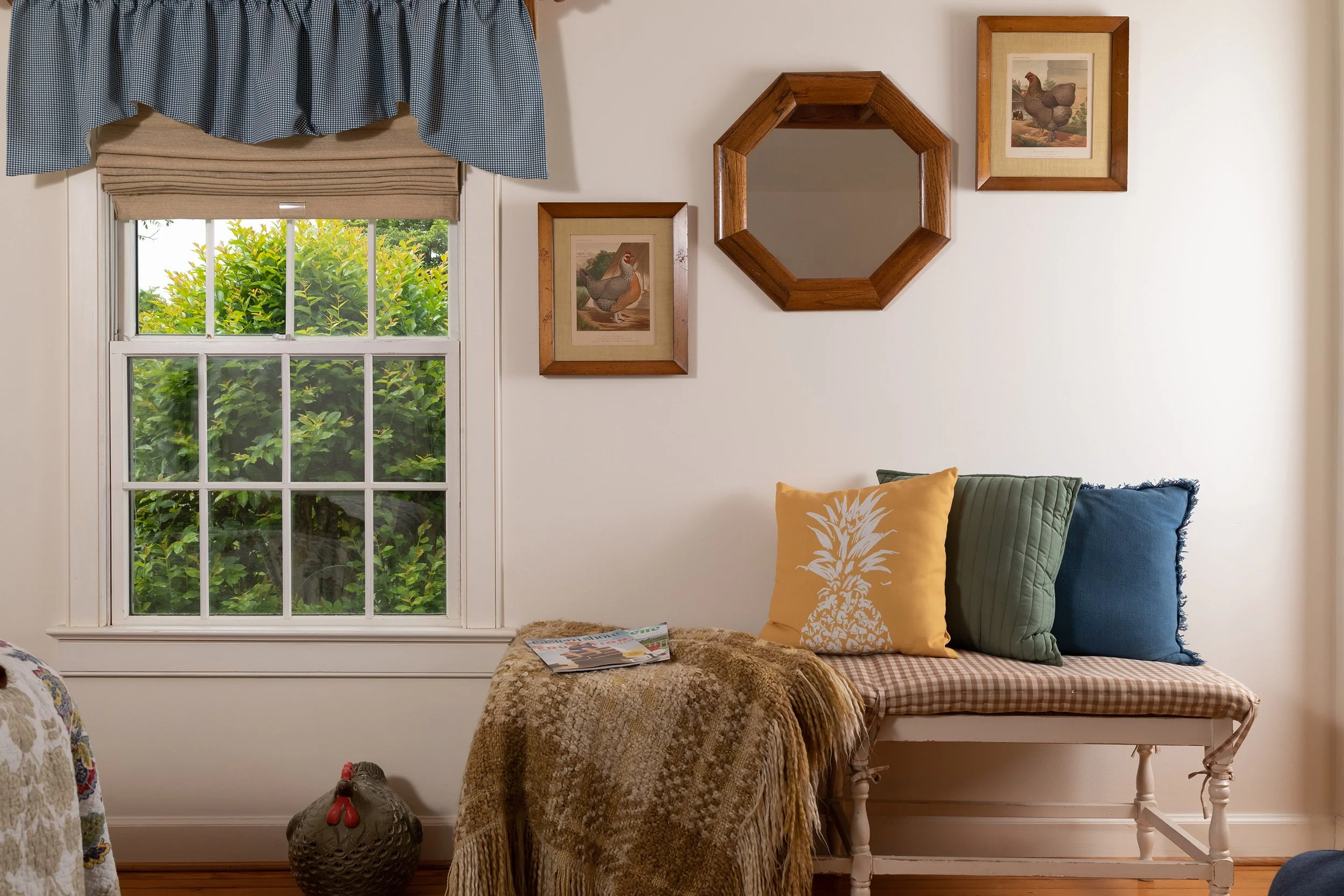 A cozy living room corner with a window showing greenery outside, framed chicken artwork on the wall, a octagonal mirror, a wooden bench with three pillows in yellow, green, and blue, a beige checkered cushion, a brown and beige throw blanket, a maga