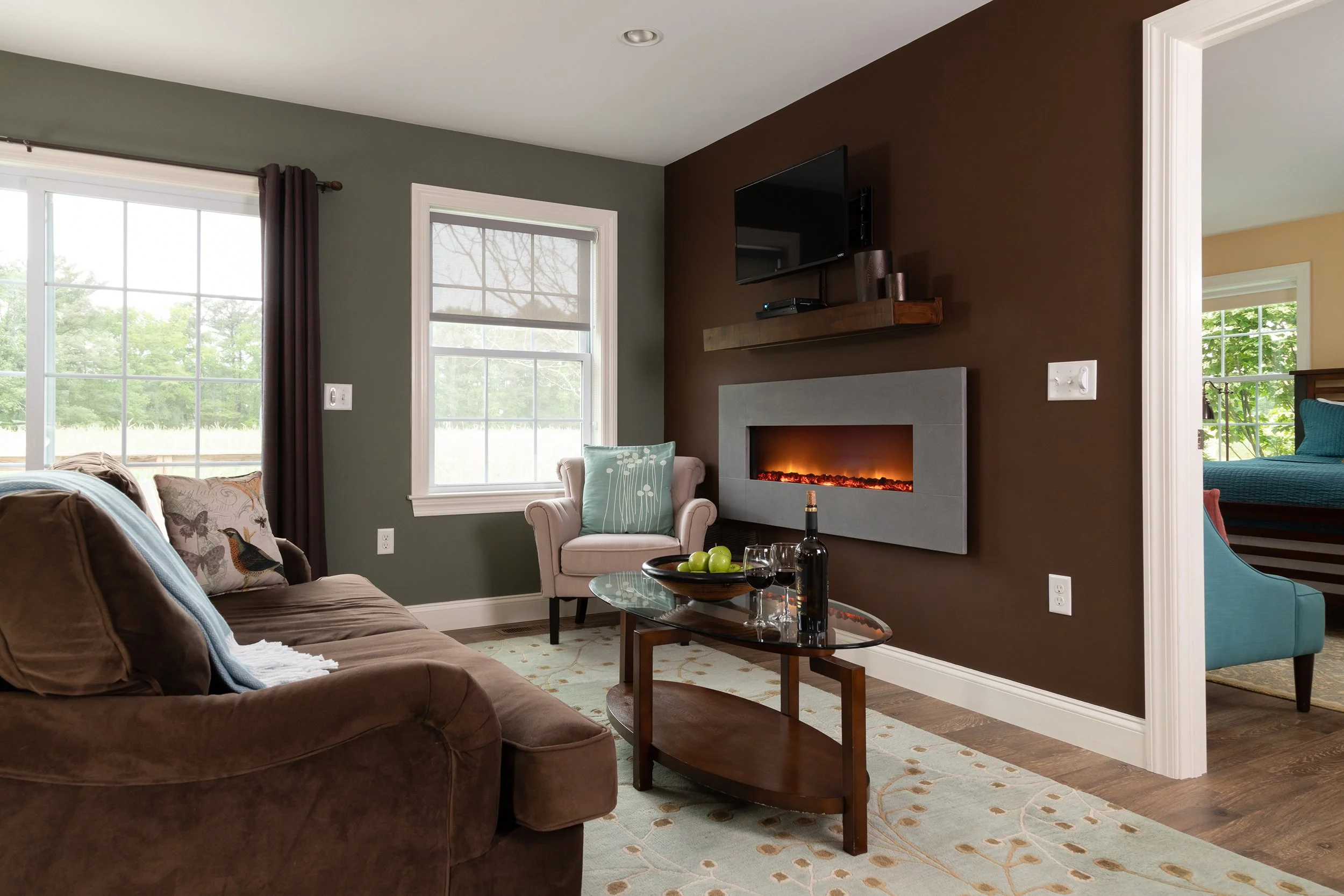 Living room with green and brown walls, a brown sofa, light-colored armchair, glass coffee table with wine and fruit, mounted flat-screen TV, and electric fireplace, with large windows showing an outdoor view.