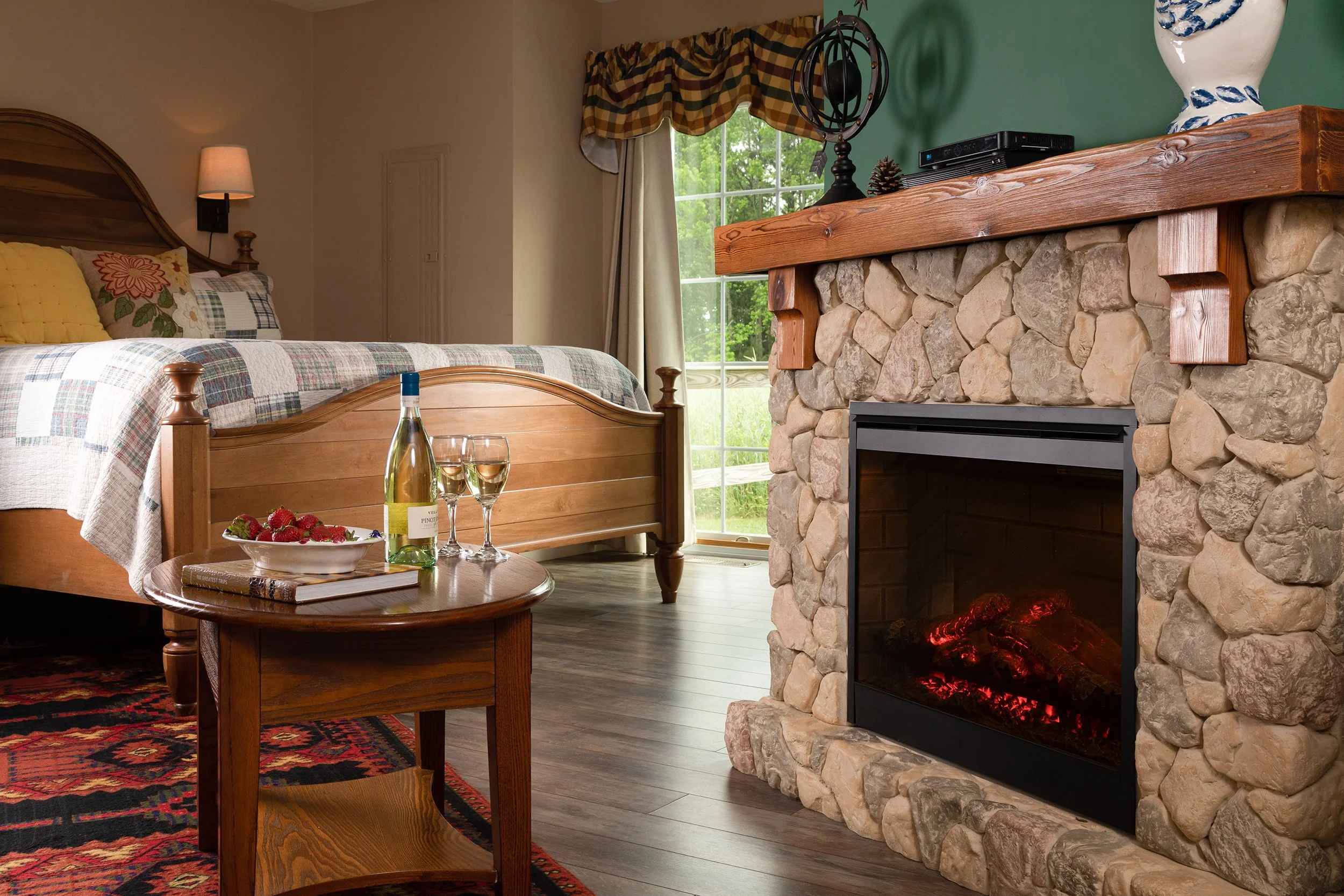 A cozy bedroom featuring a wooden bed with a plaid bedspread and pillows, a small wooden table with a bowl of strawberries, a bottle of white wine, and two glasses of wine. There's a stone fireplace with a wooden mantel and a large window with curtai