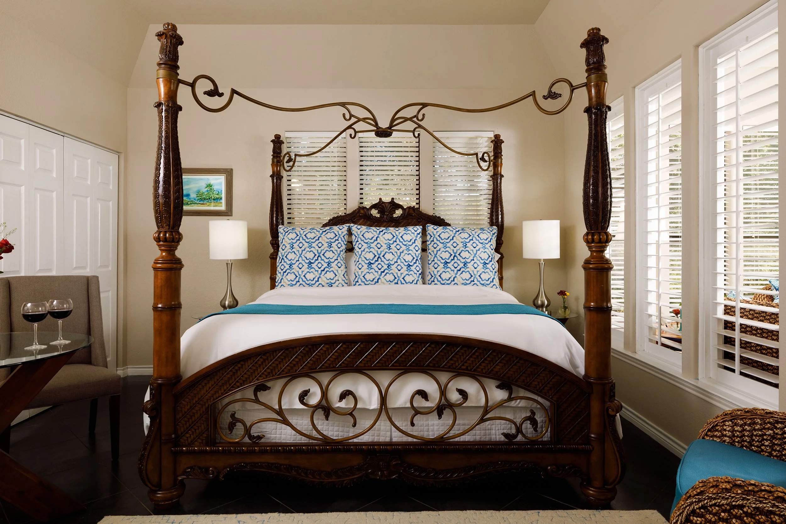 A luxurious bedroom featuring a dark wooden four-poster bed with intricate carvings and metal scrollwork, four blue and white patterned pillows, white bedding, two white bedside lamps, a small side table with a flower vase, a sitting area with wine g