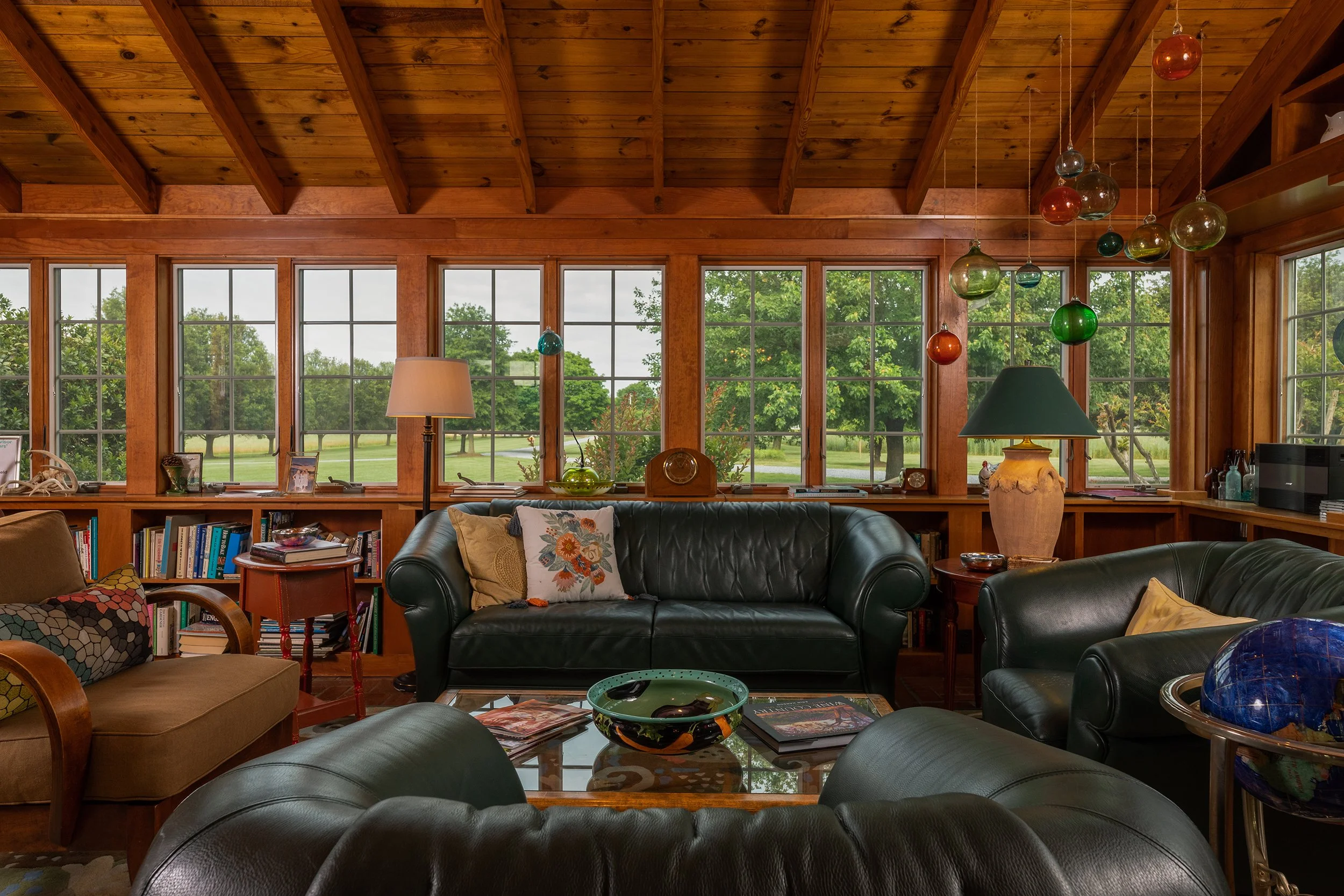 Cozy living room with large windows overlooking a green outdoor landscape, wood-paneled ceiling, leather sofas, colorful pendant lights, bookshelves, lamps, and decorative items.