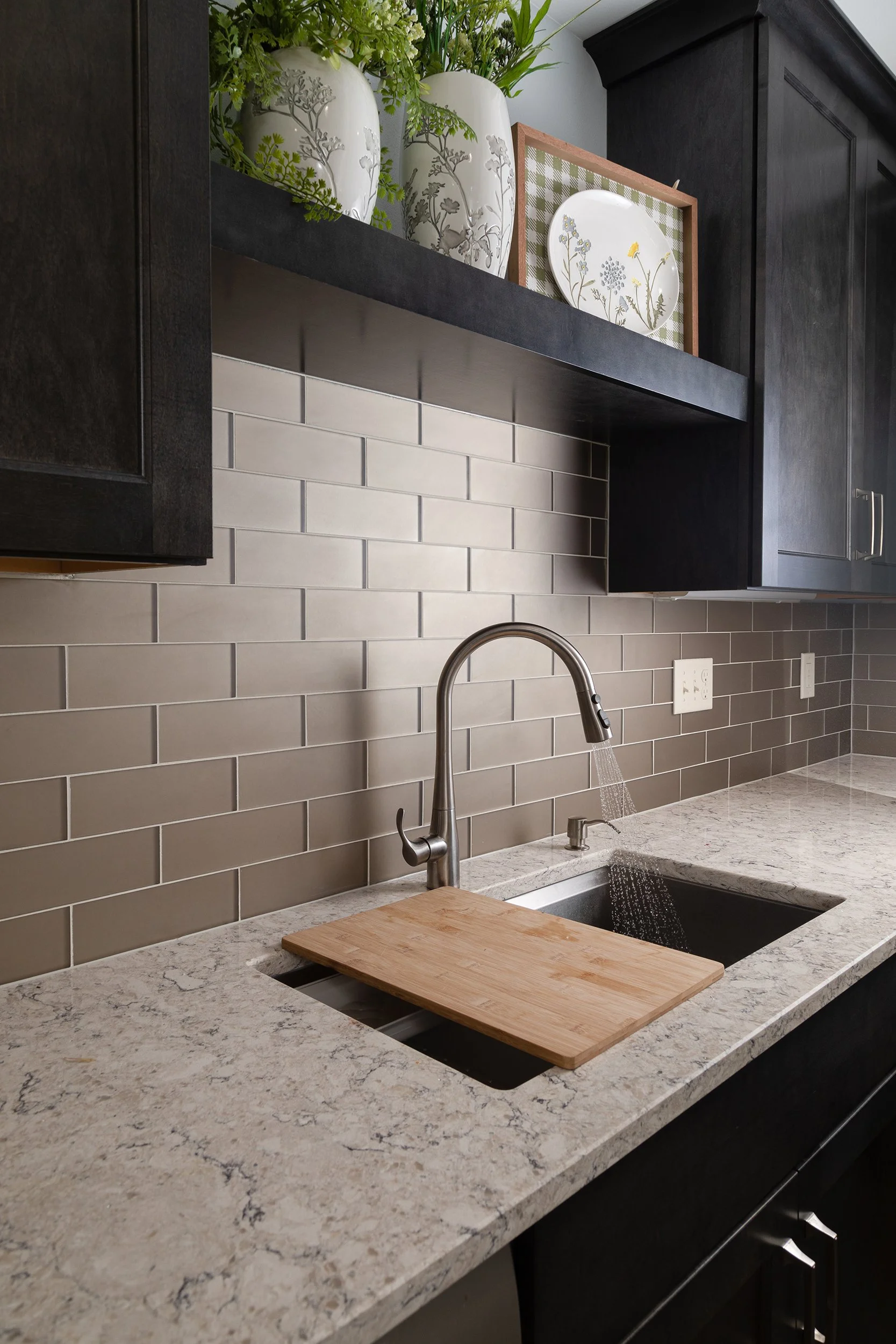 A kitchen countertop with a stainless steel sink, a wooden cutting board, beige subway tile backsplash, dark cabinetry, and decorative plates and vases with greenery on a shelf above.