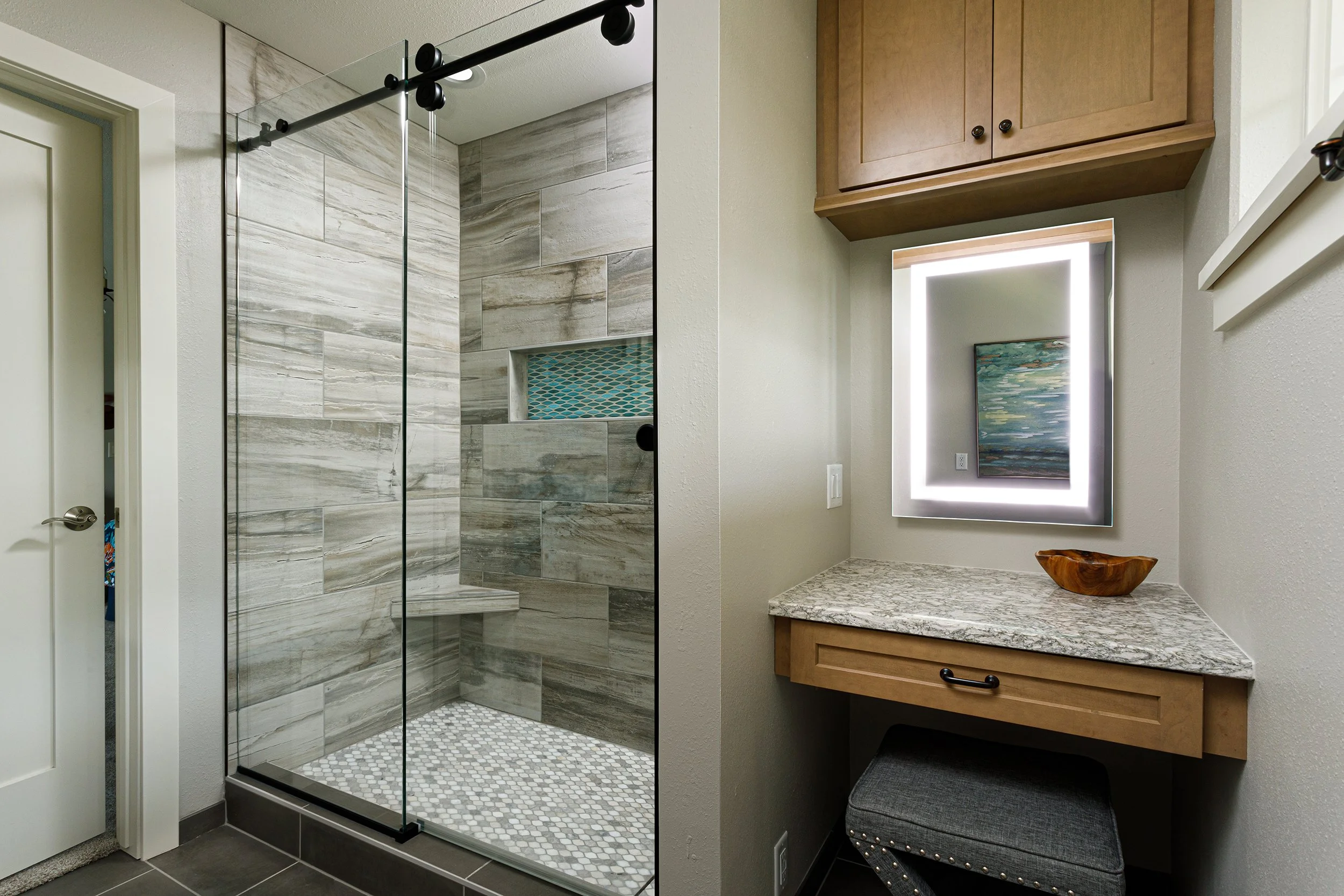 Bathroom with a walk-in glass shower, wood cabinets, vanity with granite countertop, framed mirror with lighting, and a small stool.