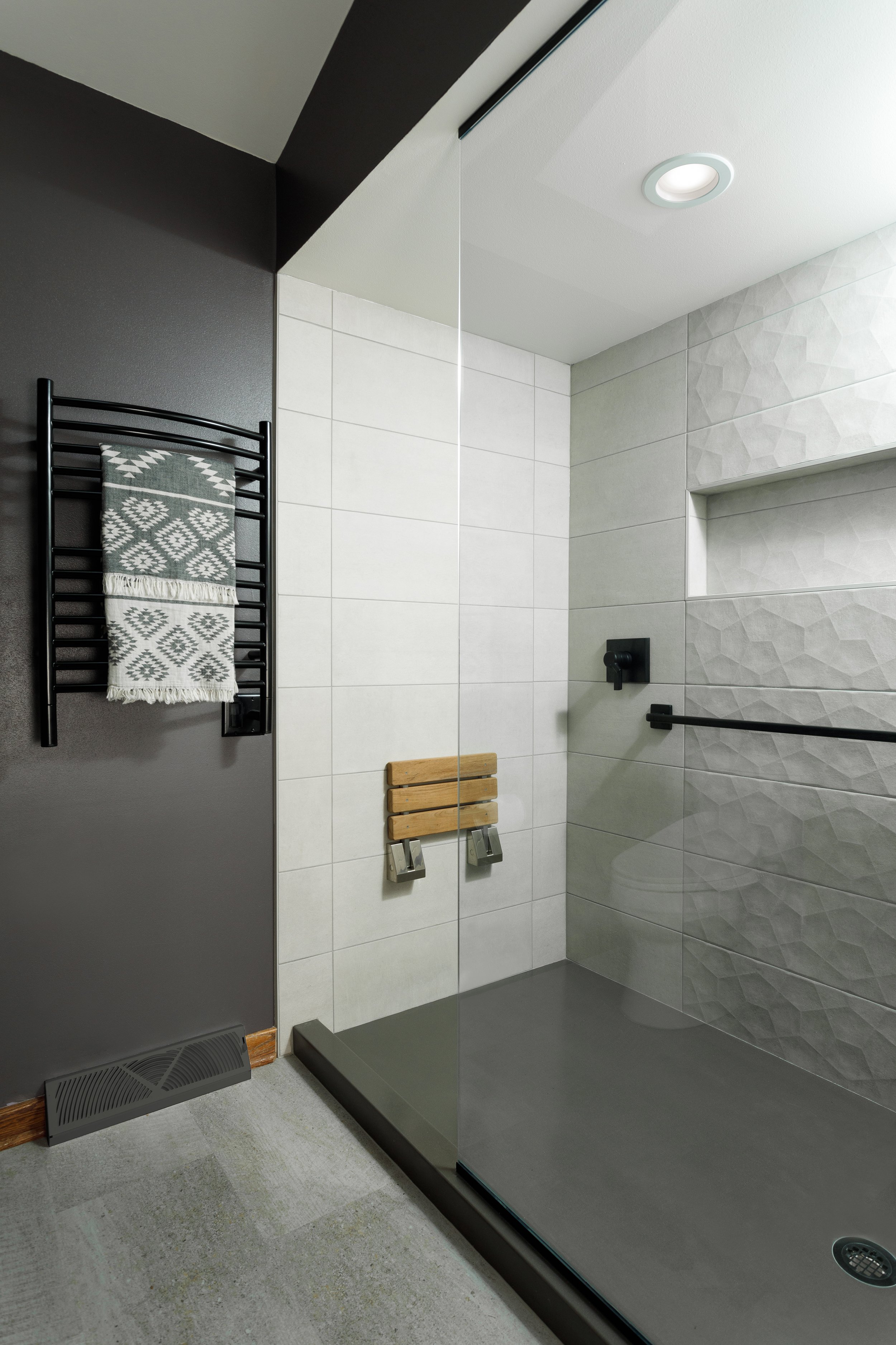 A modern shower area with a glass enclosure, white textured tiles, a wooden bench, black fixtures, a black towel rack with a patterned towel, and a grey floor.