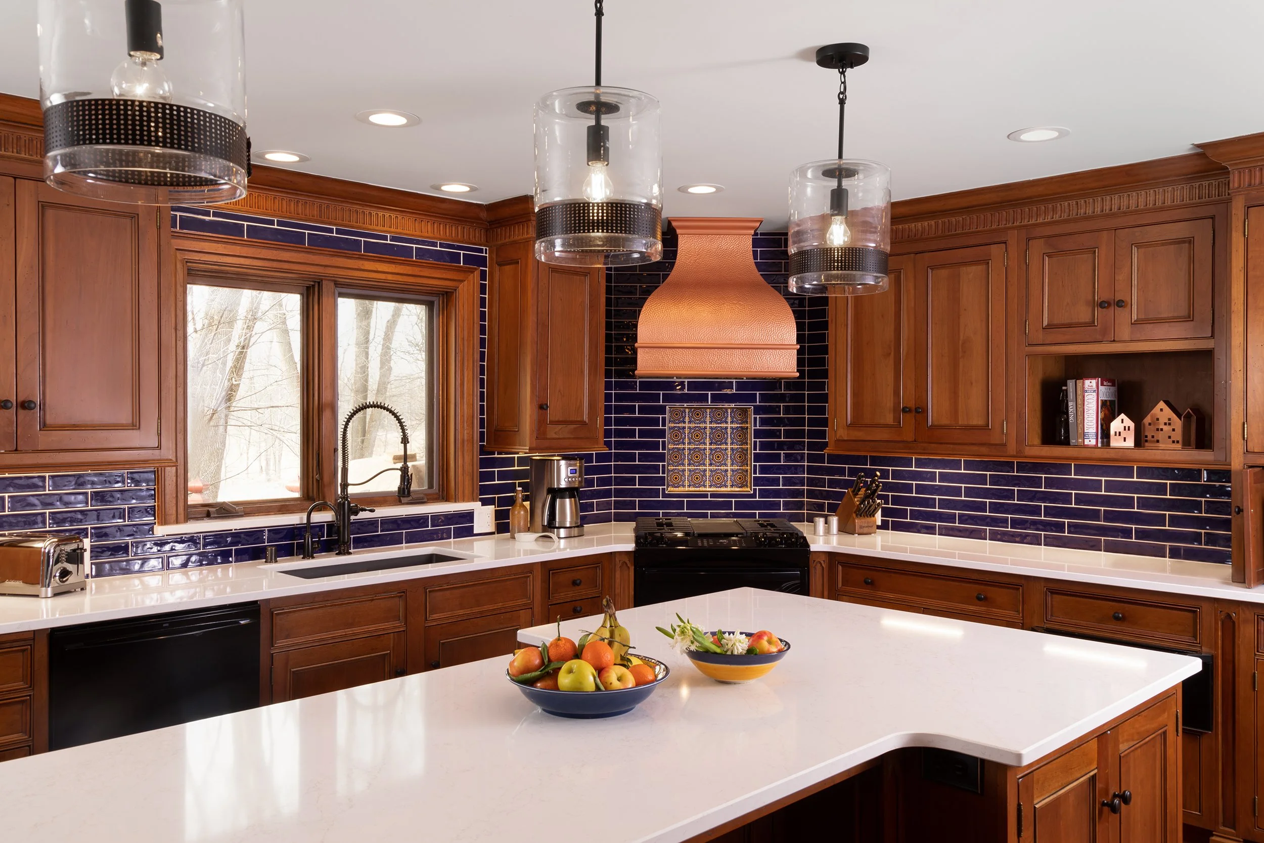 Kitchen with wooden cabinets, navy blue tile backsplash, white countertops, and a large island with fruit bowls. There are three pendant lights hanging above the island and a window over the sink showing a wintery outdoor scene.