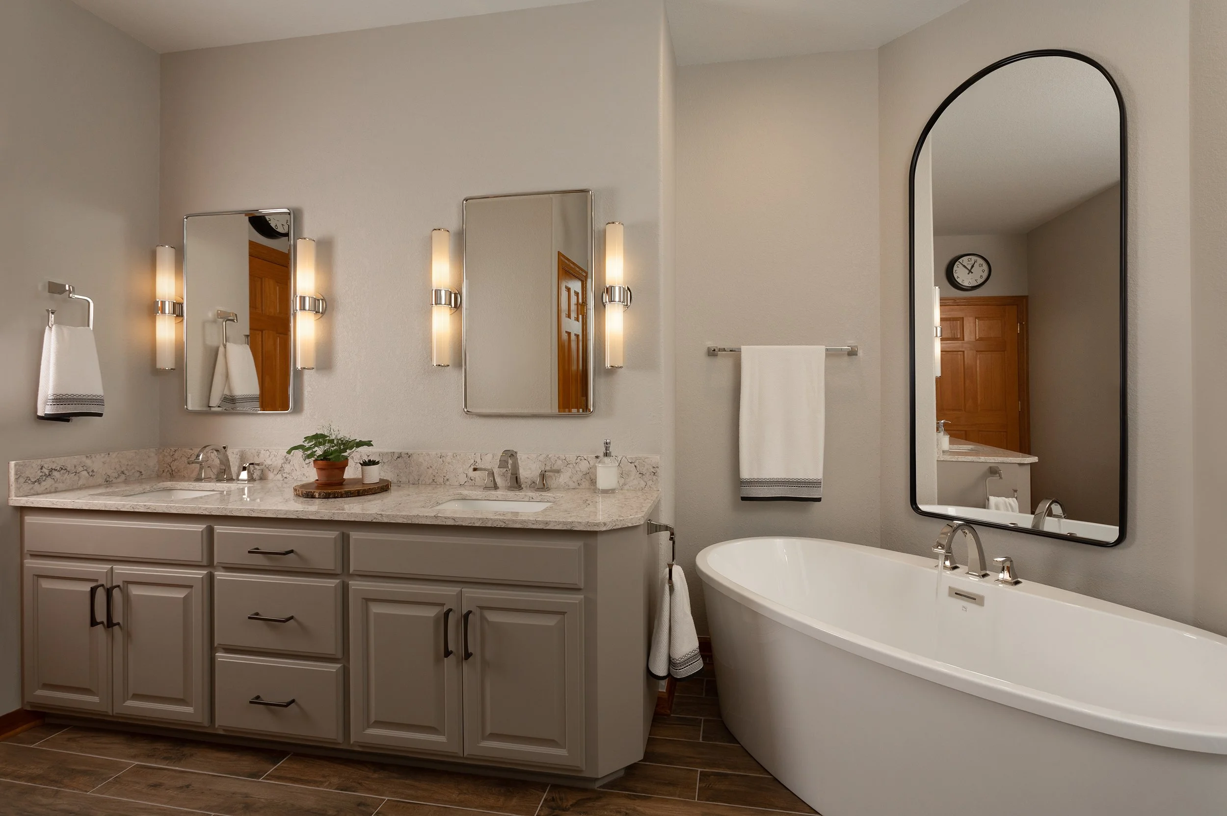 Modern bathroom with double sink vanity, two mirrors, wall-mounted lights, a freestanding bathtub and a large mirror reflecting part of the room, including a clock and wooden door.