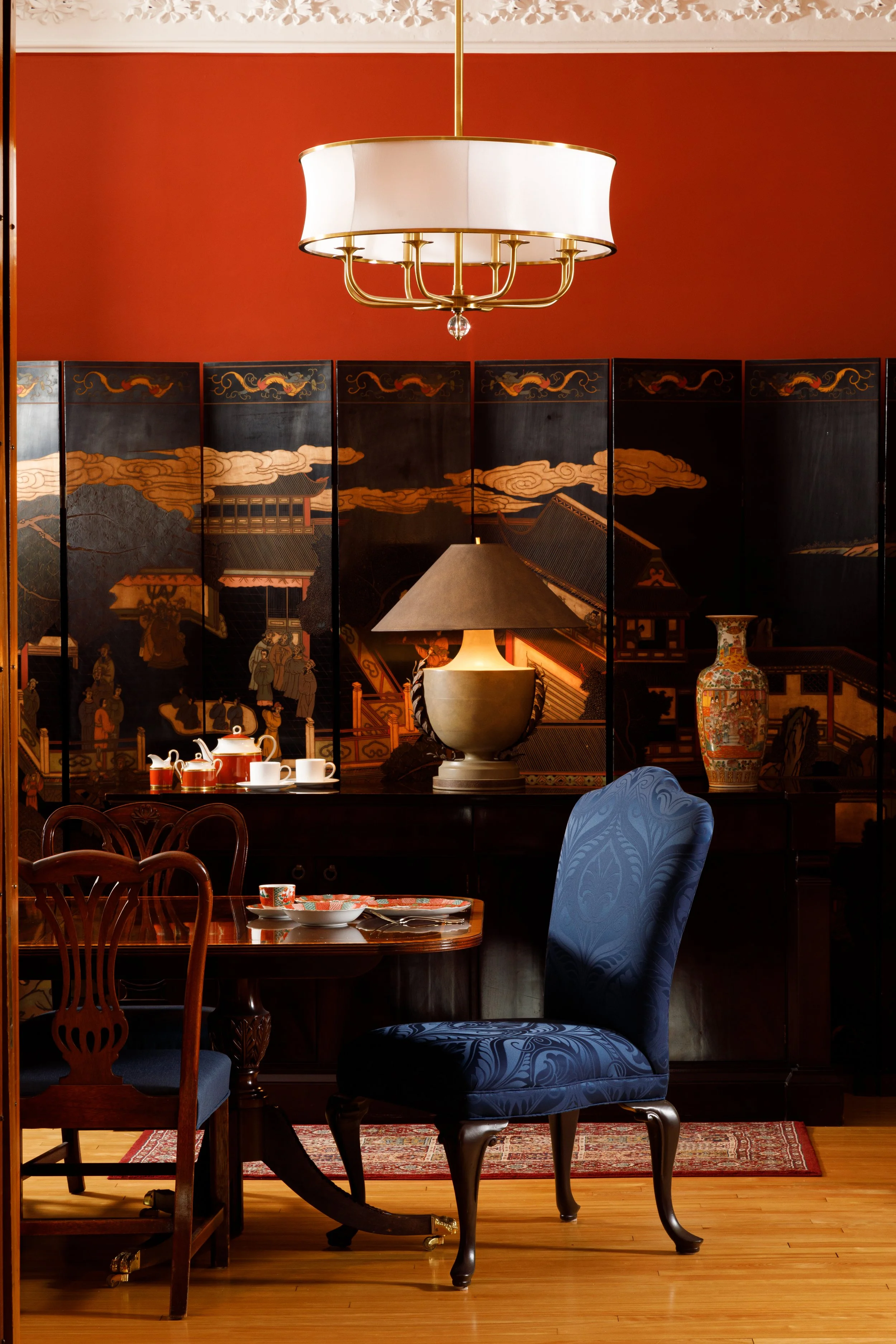 A dining room with a dark wooden table and chairs, a large blue upholstered chair, a decorative screen with traditional Asian artwork, a table lamp, a floral vase, and a chandelier hanging from the ceiling.