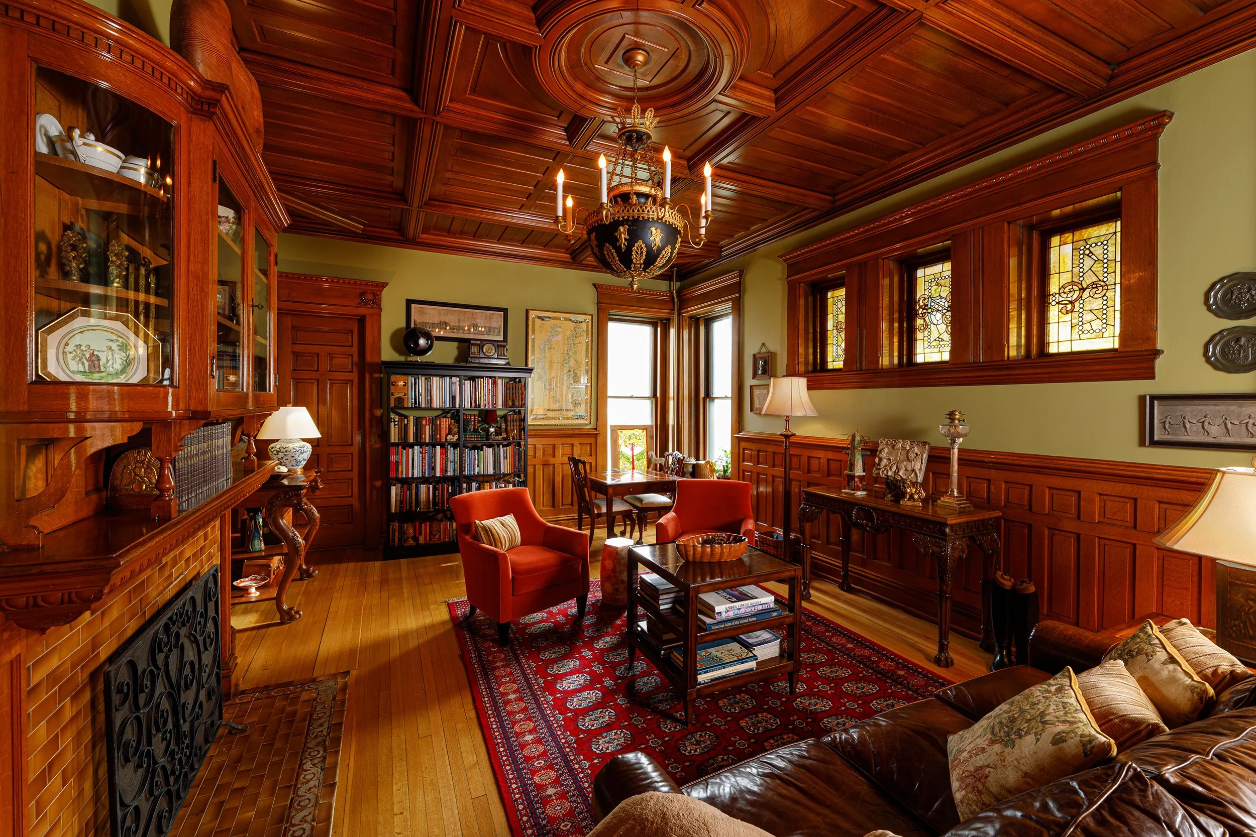 A traditional living room with wood-paneled walls, a wooden ceiling, a chandelier, a leather sofa with pillows, an area rug, armchairs, a coffee table, a bookshelf, and decorative stained glass windows.