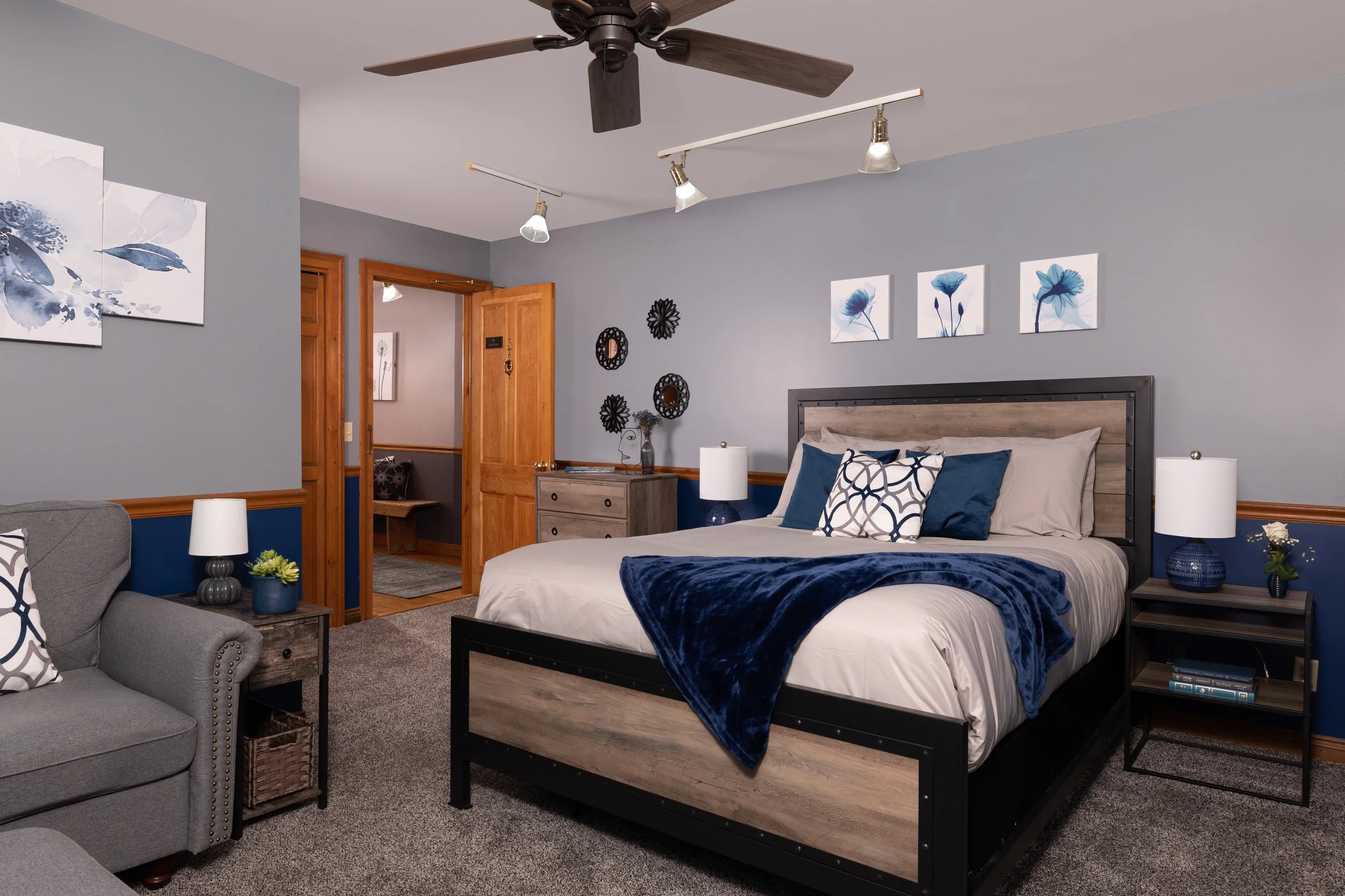 A modern bedroom with a gray wall, wooden furniture, a bed with a wooden headboard, blue and beige bedding, and artwork of blue flowers.