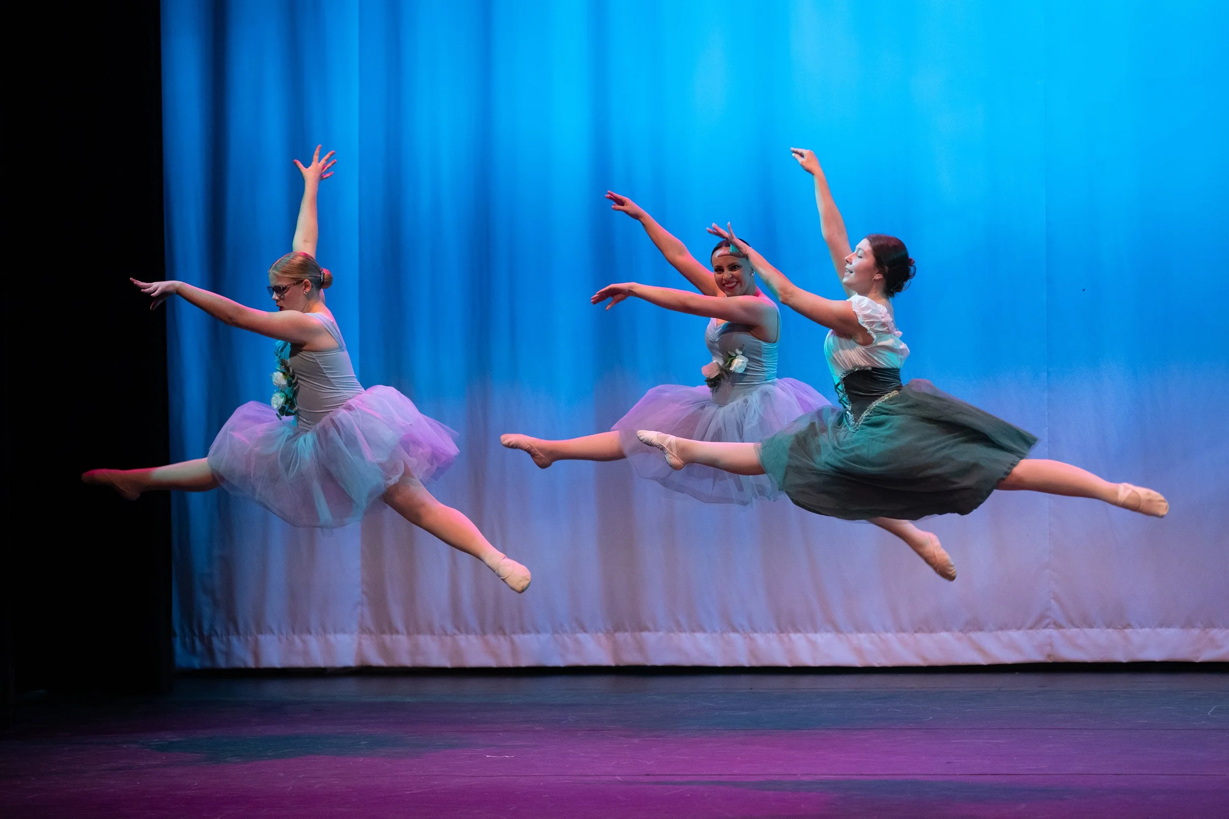 Three ballet dancers in tutus performing a leap on stage with a blue curtain background.
