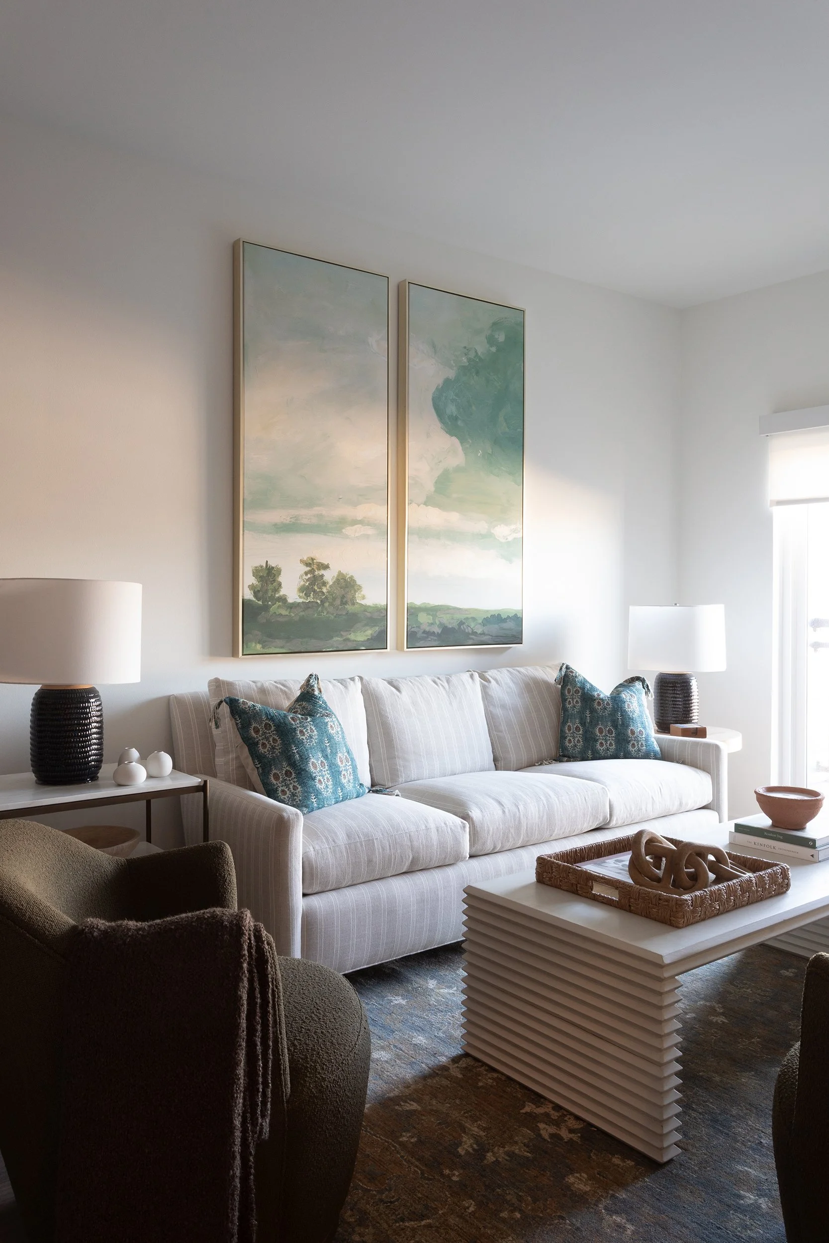 Living room with a white striped sofa, two blue patterned throw pillows, two table lamps on side tables, abstract landscape artwork above the sofa, a coffee table with decorative items, and a window with white blinds.