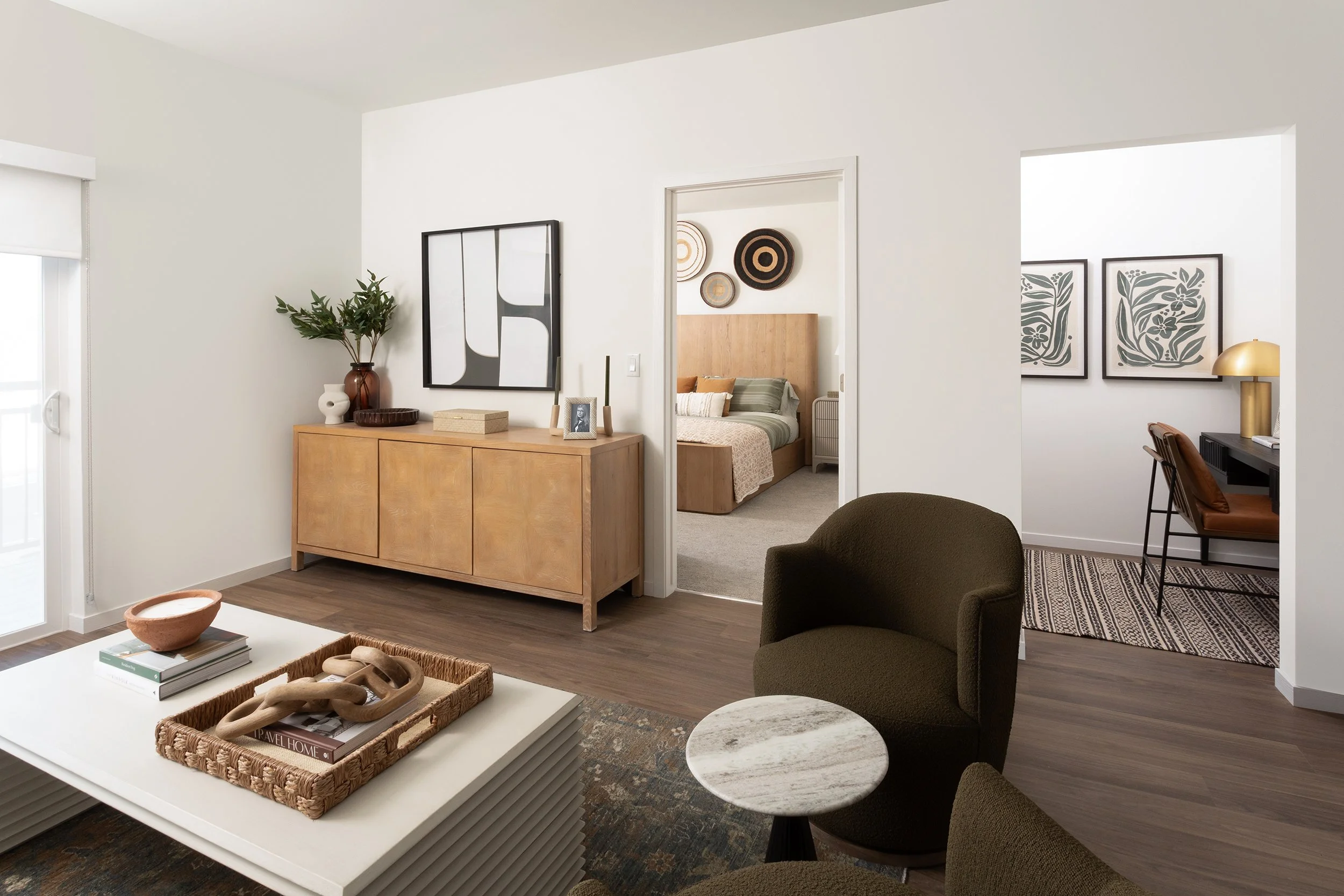 Living room with a wooden sideboard, green armchair, coffee table with decorative objects, and a view of a bedroom and a small workspace in the background.